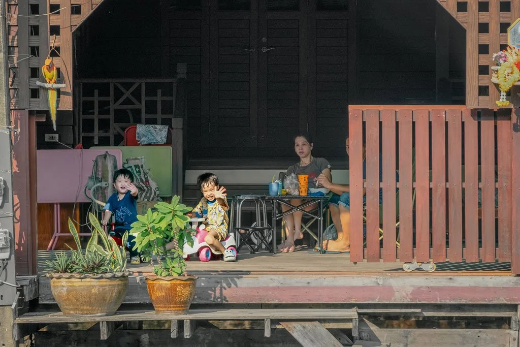 Two children playing on a porch with a woman sitting at a table, surrounded by potted plants and household items.