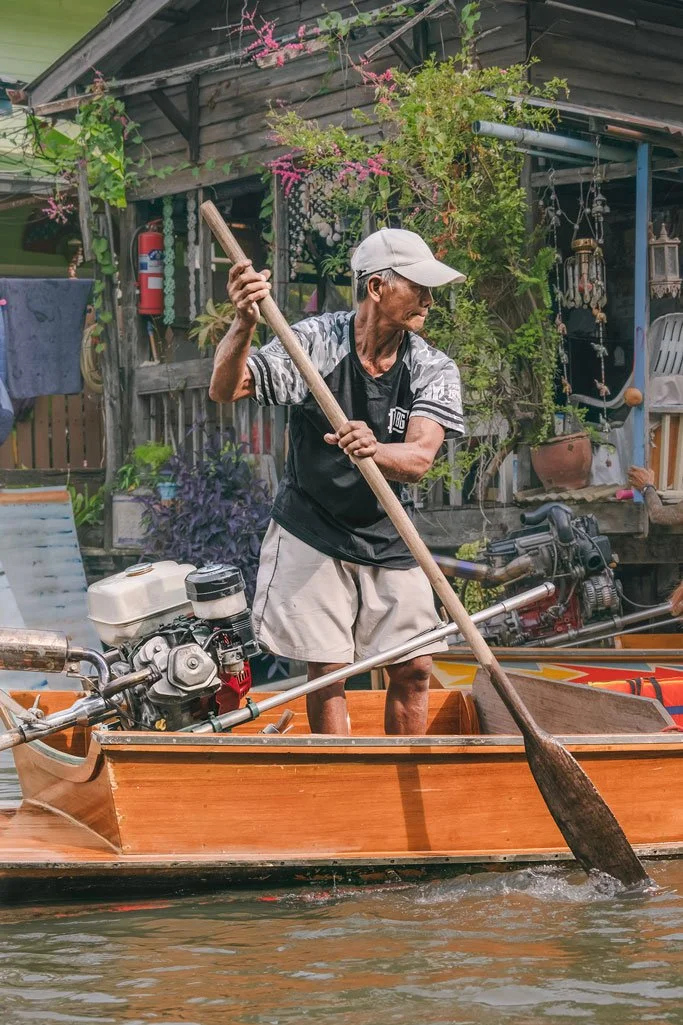 A man in a baseball cap and casual clothing paddling a wooden boat along a waterway with a motor attached to the back, in front of a rustic house with plants and hanging decorations.