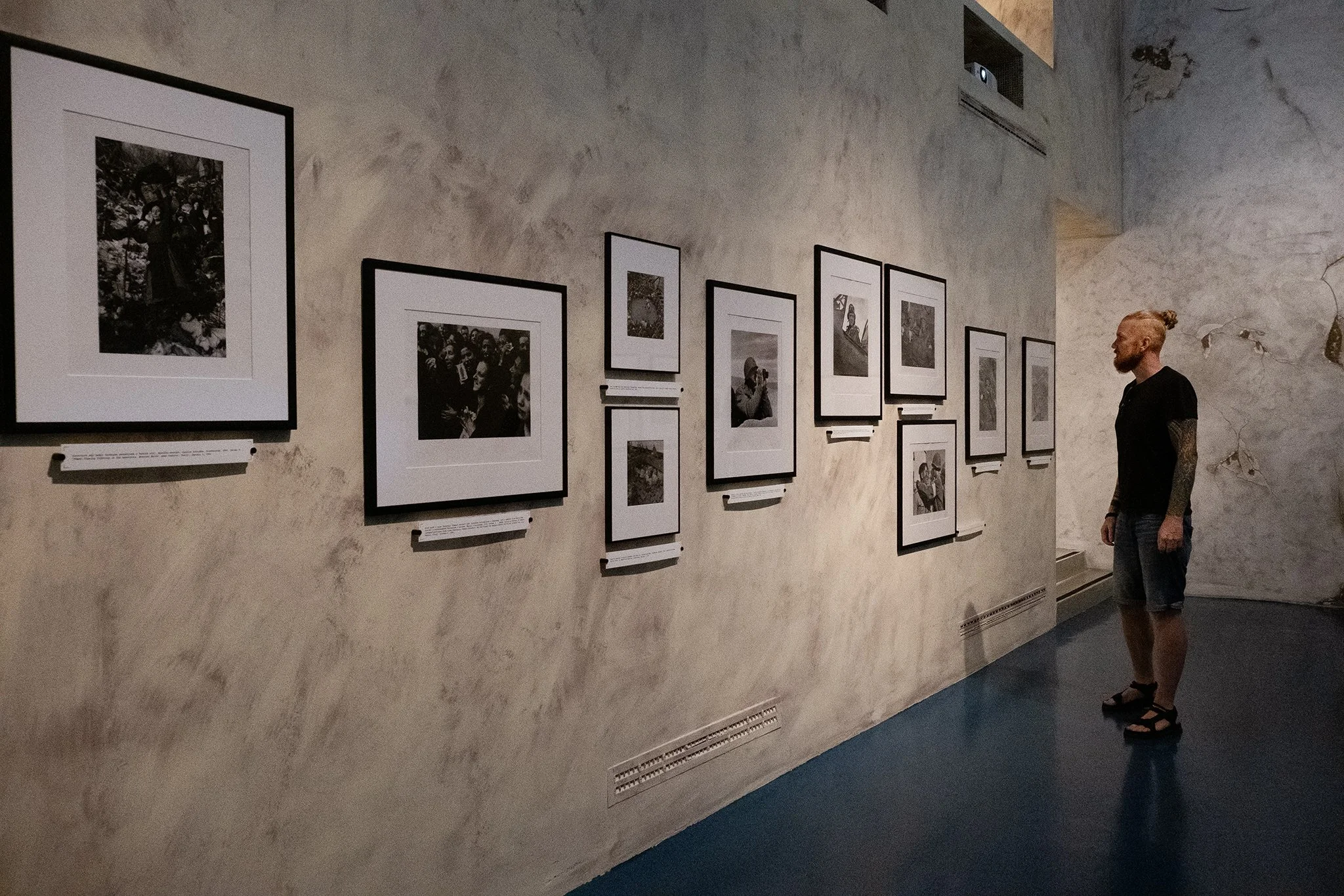 A man with blond hair and tattoos on his arm looking at black-and-white photographs on a gallery wall with information plaques underneath, in a dimly lit art gallery.