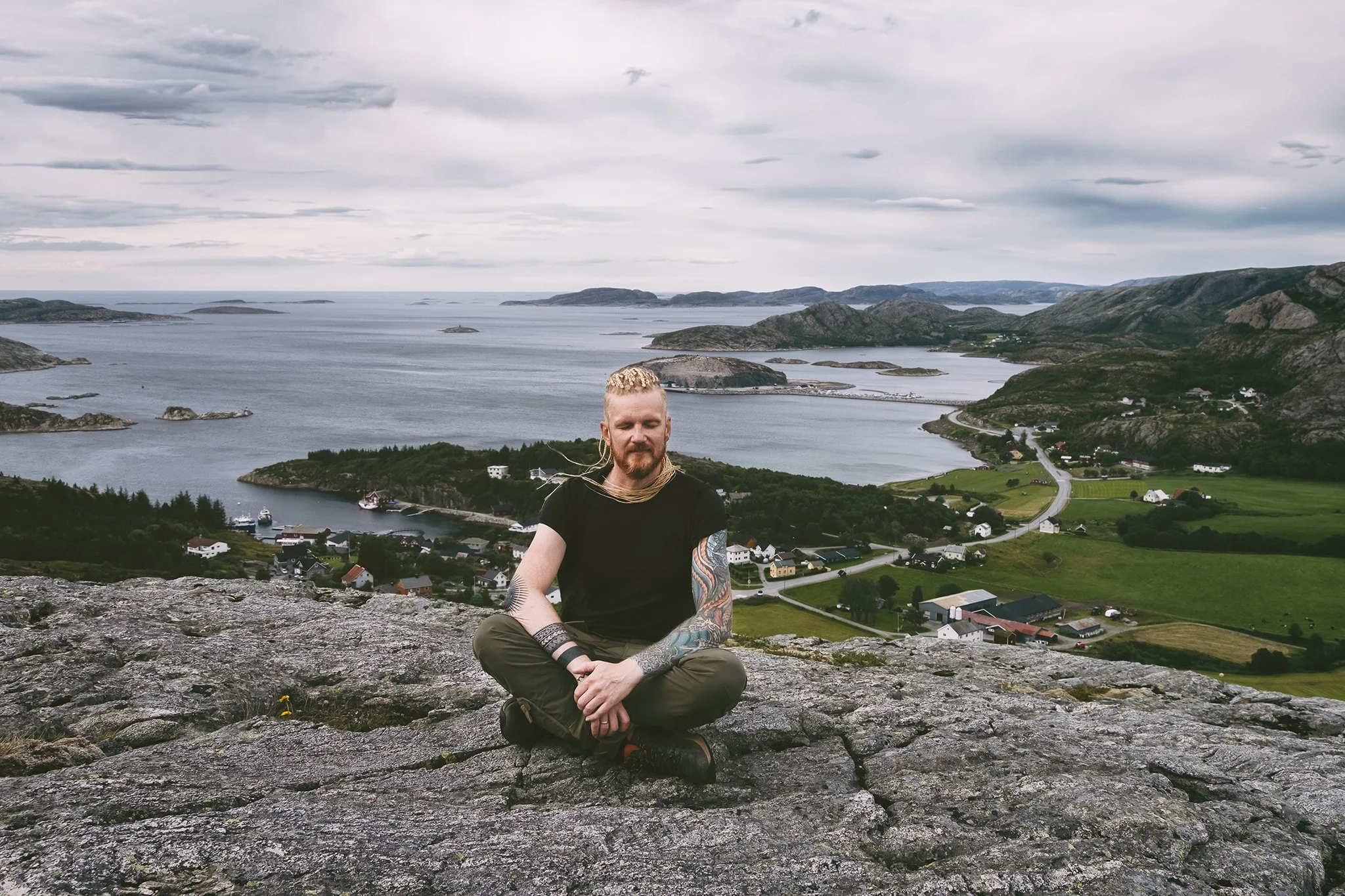A man with tattoos sitting cross-legged on rocks, overlooking a scenic coastline with islands, water, and houses under a cloudy sky.