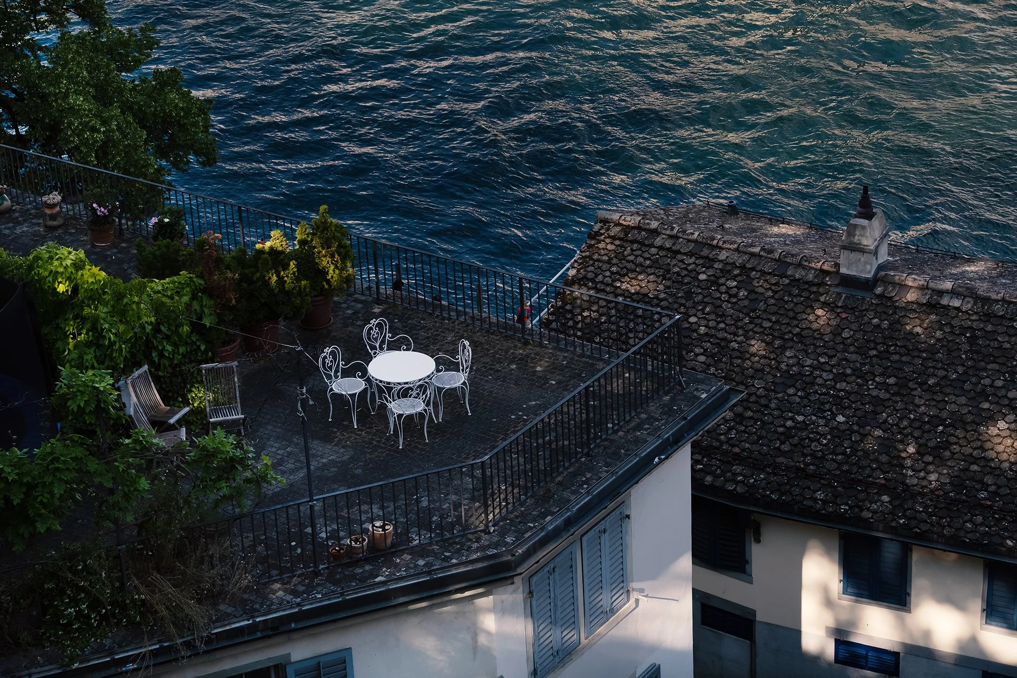 A rooftop terrace overlooking a body of water, with potted plants, a white table and chairs, and a stone chimney on the roof.