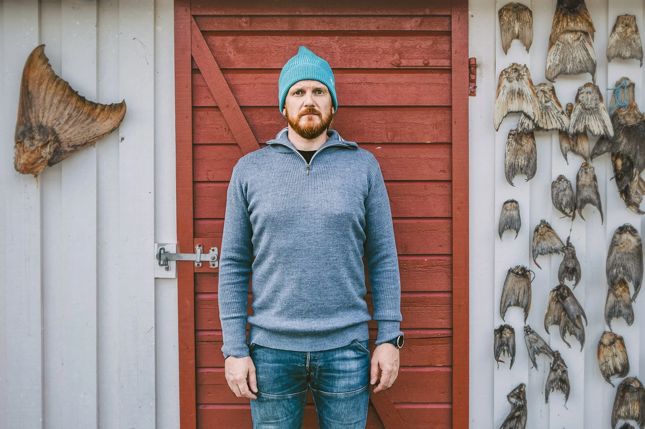 A man wearing a blue knit cap, a grey zip-up sweater, and jeans stands in front of a red wooden door with a grey and white gradient wall on either side. On the right, dried fish are mounted on the wall, and on the left, part of a mounted fish tail is visible.