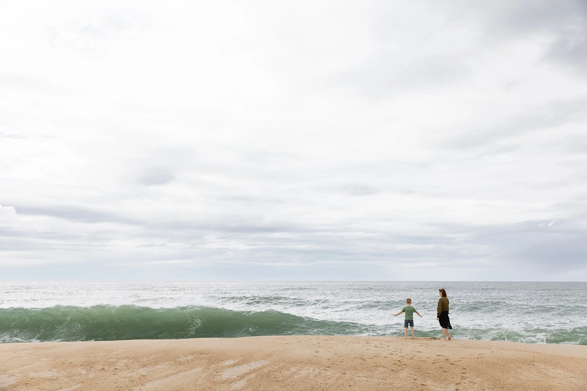Two children and an adult standing on a sandy beach near the ocean, looking at the water as waves roll in under a cloudy sky.