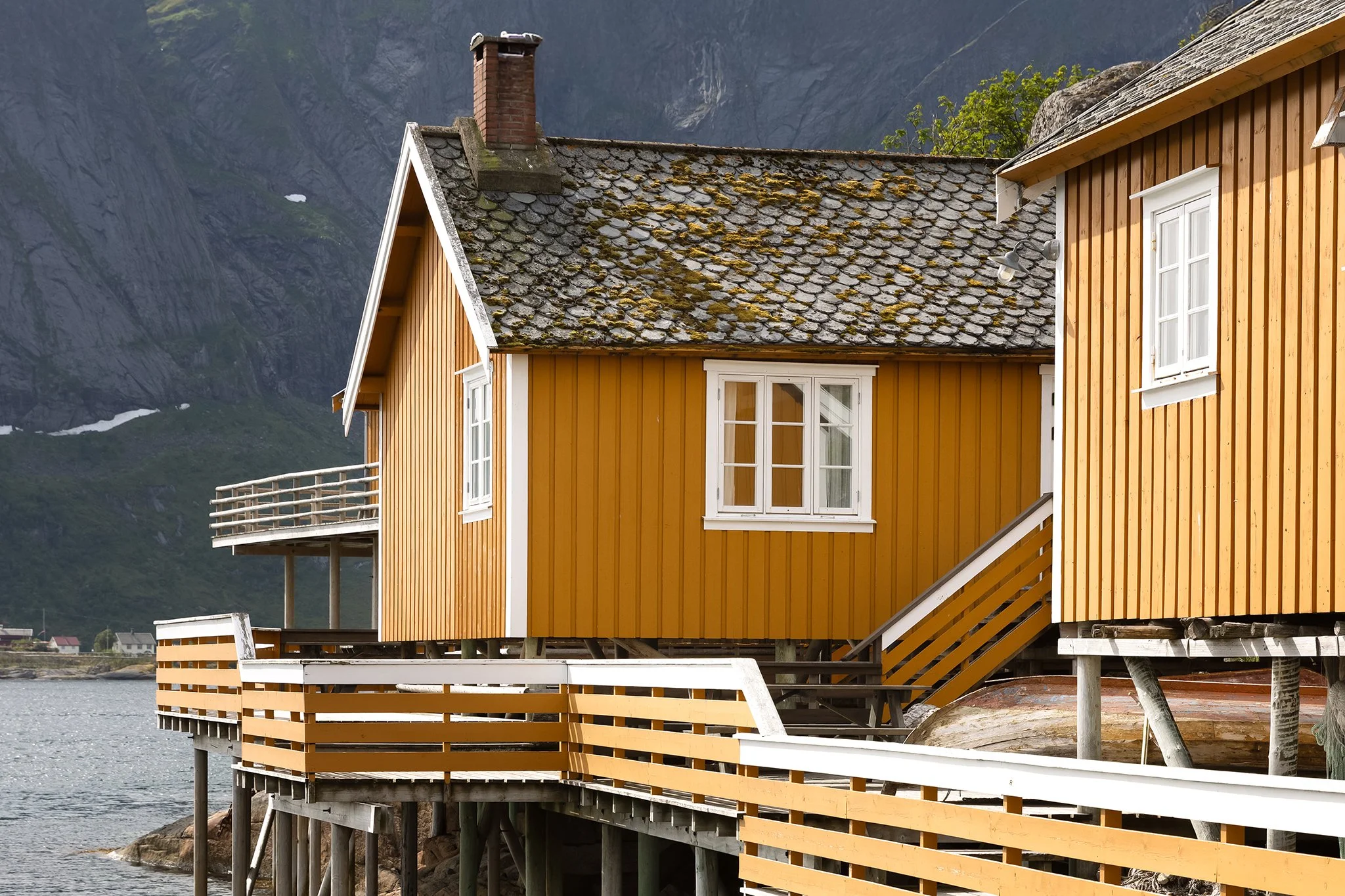 Yellow wooden house with white-framed windows and a moss-covered slate roof, built on stilts above a body of water with mountains in the background.