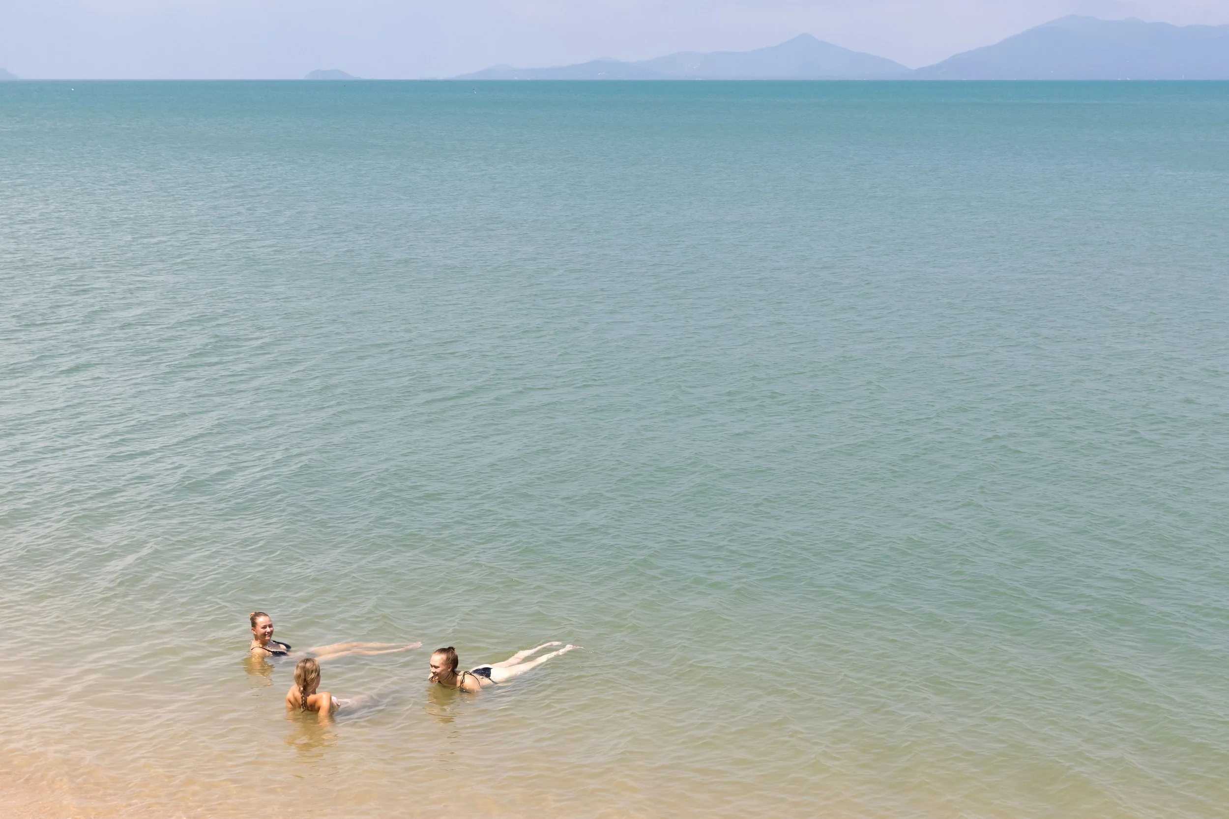 People swimming and relaxing in the ocean near the beach with distant mountains in the background.