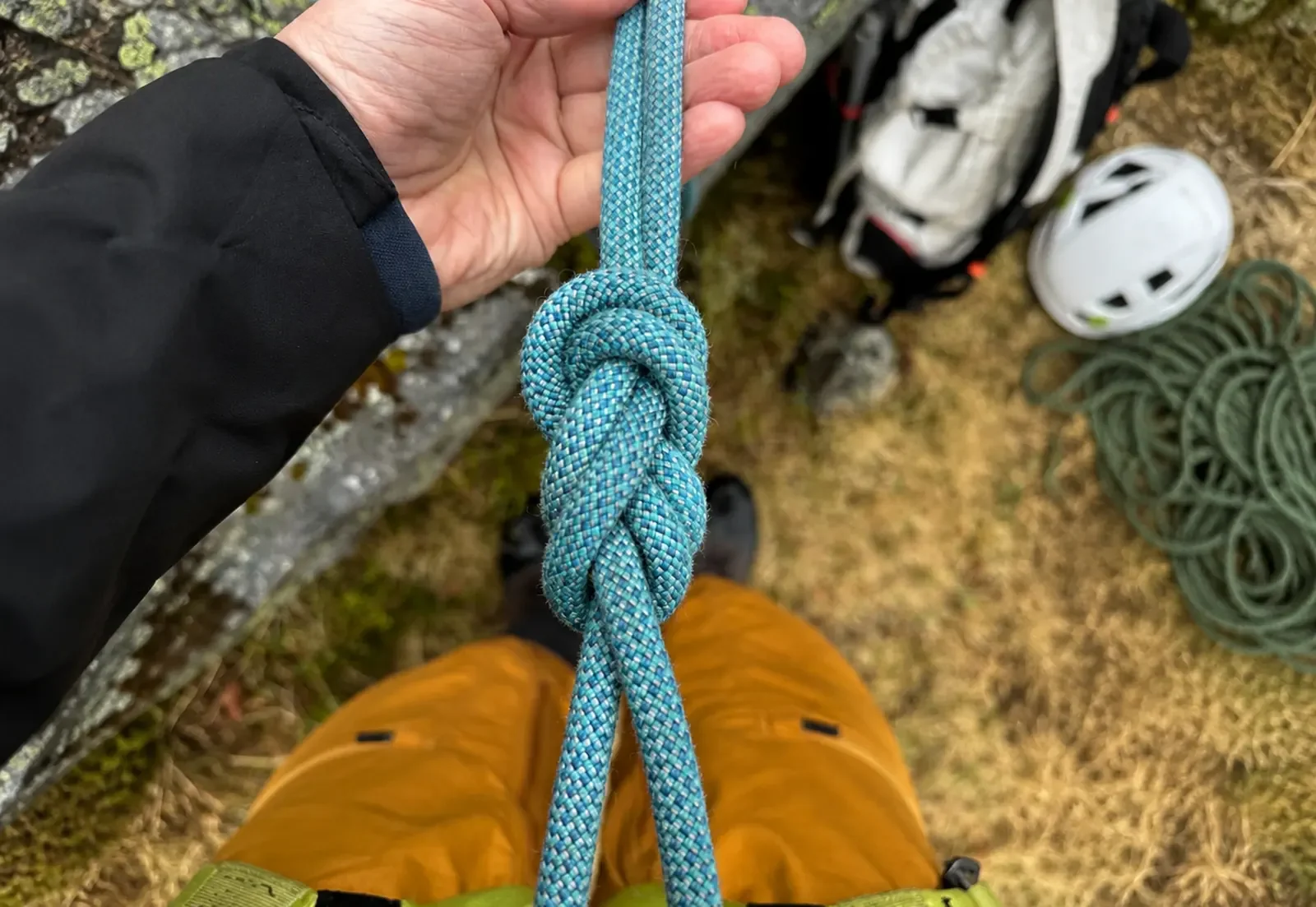 A person holding a climbing rope tied in a knot with outdoor gear and shoes on the ground below.