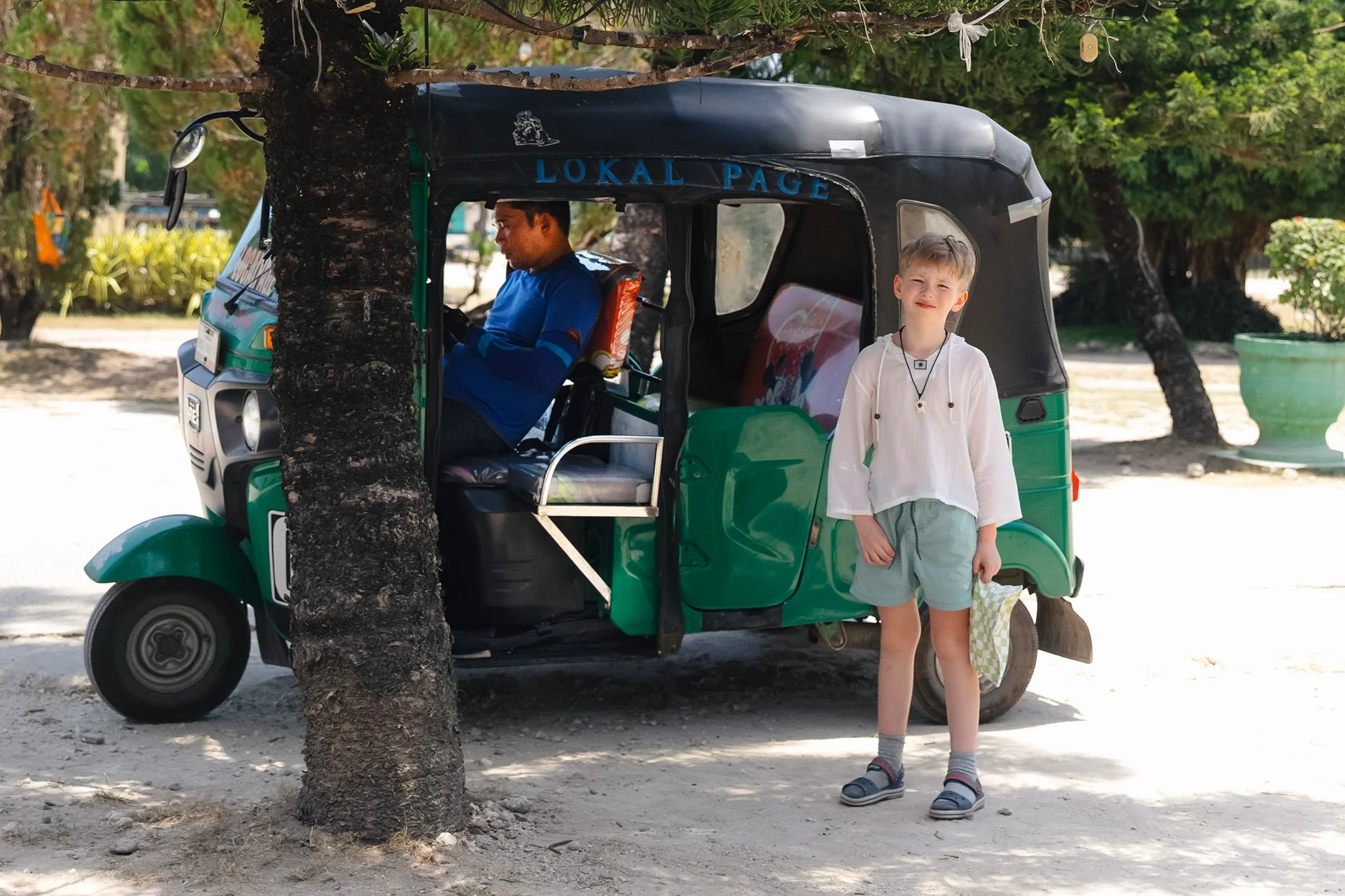 A young boy with blonde hair, wearing a white shirt, gray shorts, and sandals, stands outdoors near a green tuk-tuk with a black roof. He is looking at the camera with a slight smile. In the background, an adult man sitting inside the tuk-tuk, wearing a blue shirt, is looking at his phone. The scene includes trees and potted plants, indicating a warm, sunny day.