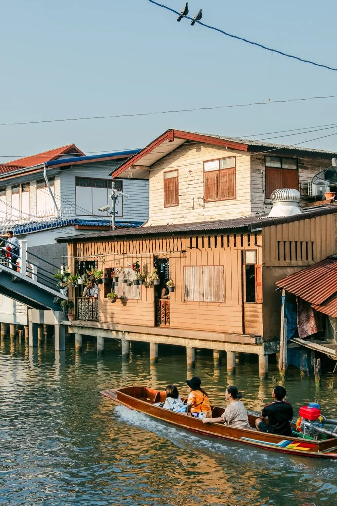 A boat with five passengers, three children and two adults, is traveling along a waterway next to stilt houses. The houses are wooden and built on the water, with some decorated with hanging plants and people standing on the balcony. Overhead, there are power lines and birds perched on a wire, with a clear sky above.