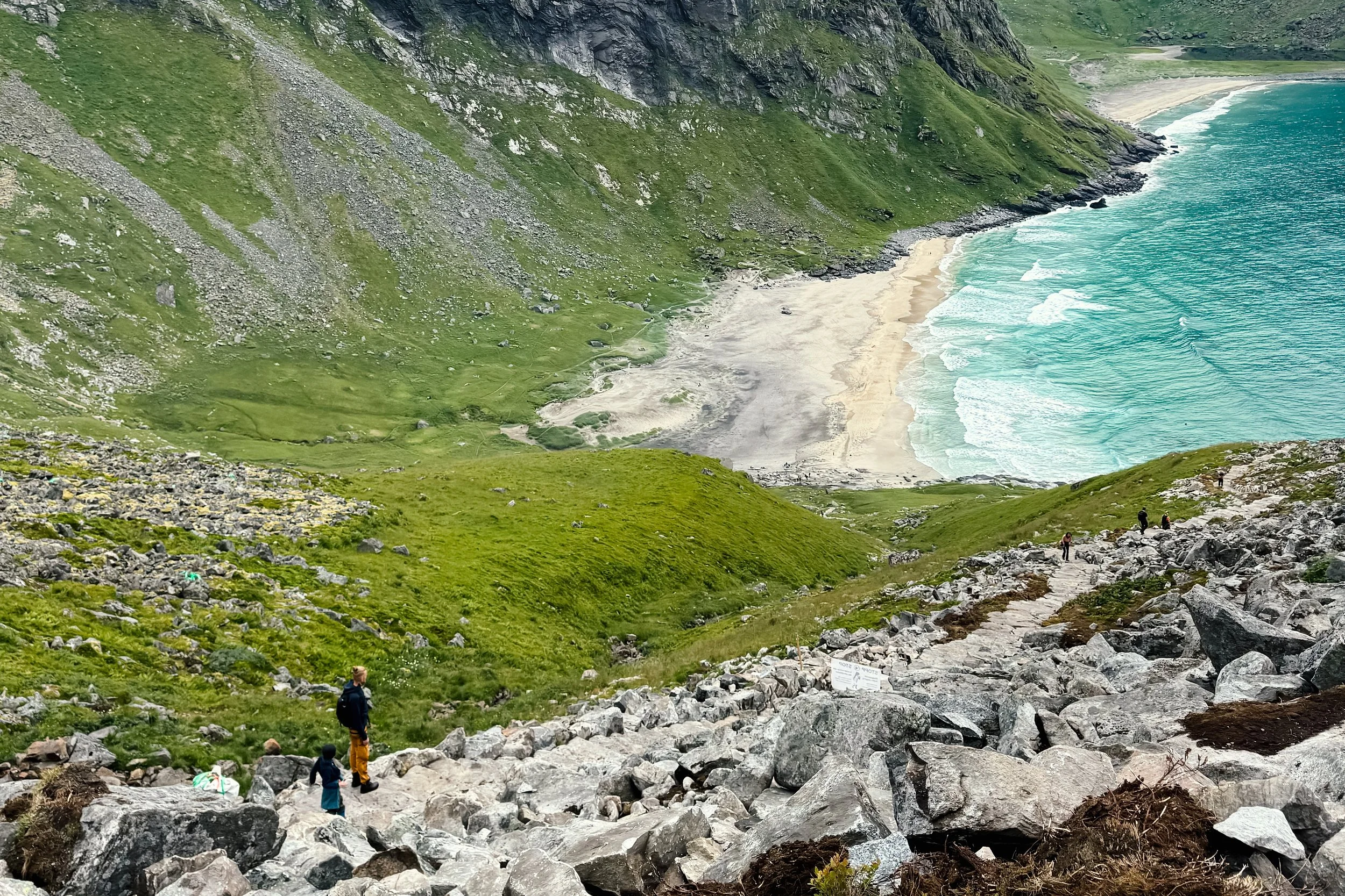 A scenic view of a mountainous coastline with a sandy beach and ocean waves. Green grassy slopes and rocky terrain are visible, with a few hikers on the trail.