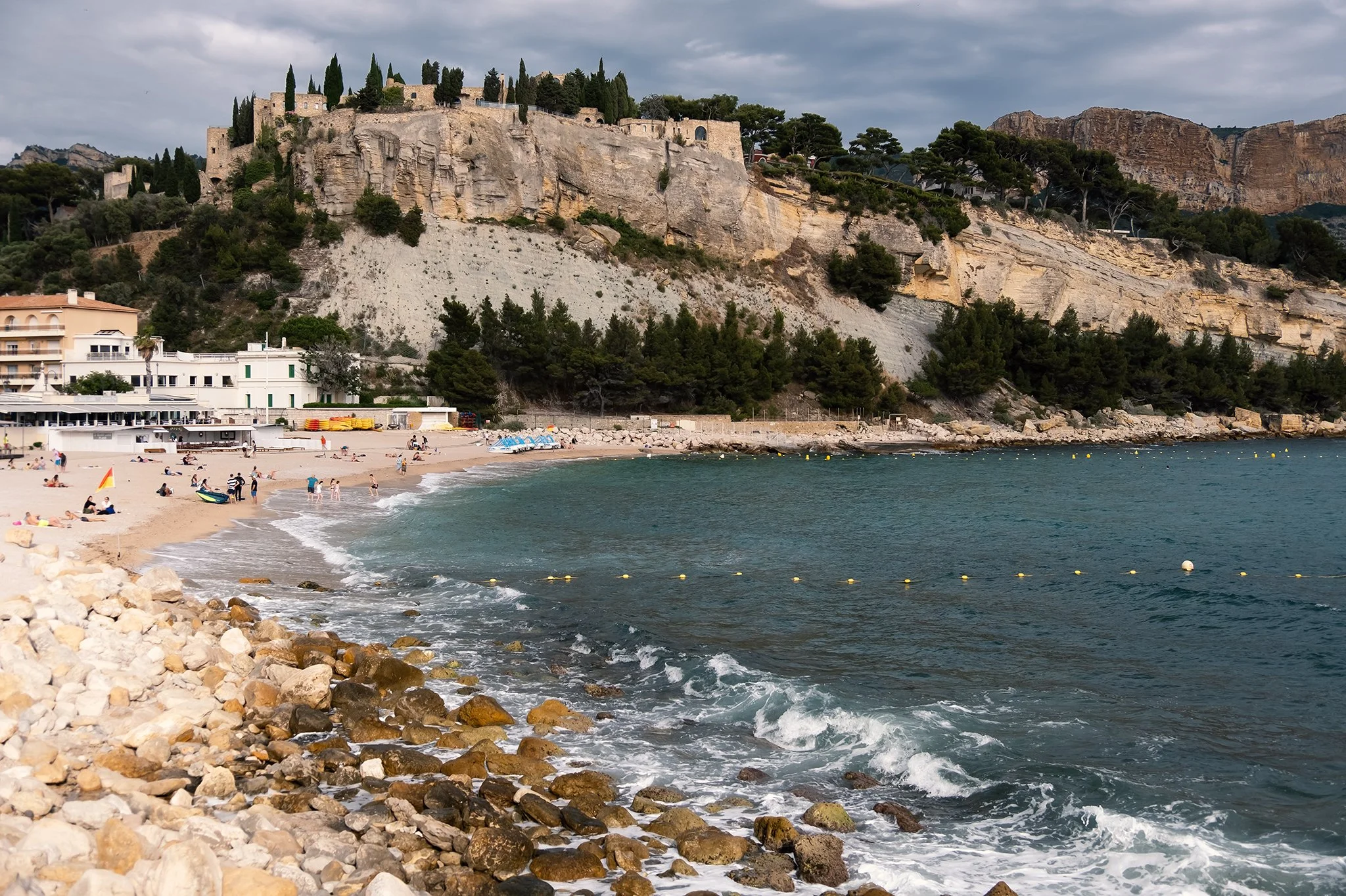 Beach with people sunbathing and swimming, rocky shoreline, hillside with trees and buildings, cloudy sky.