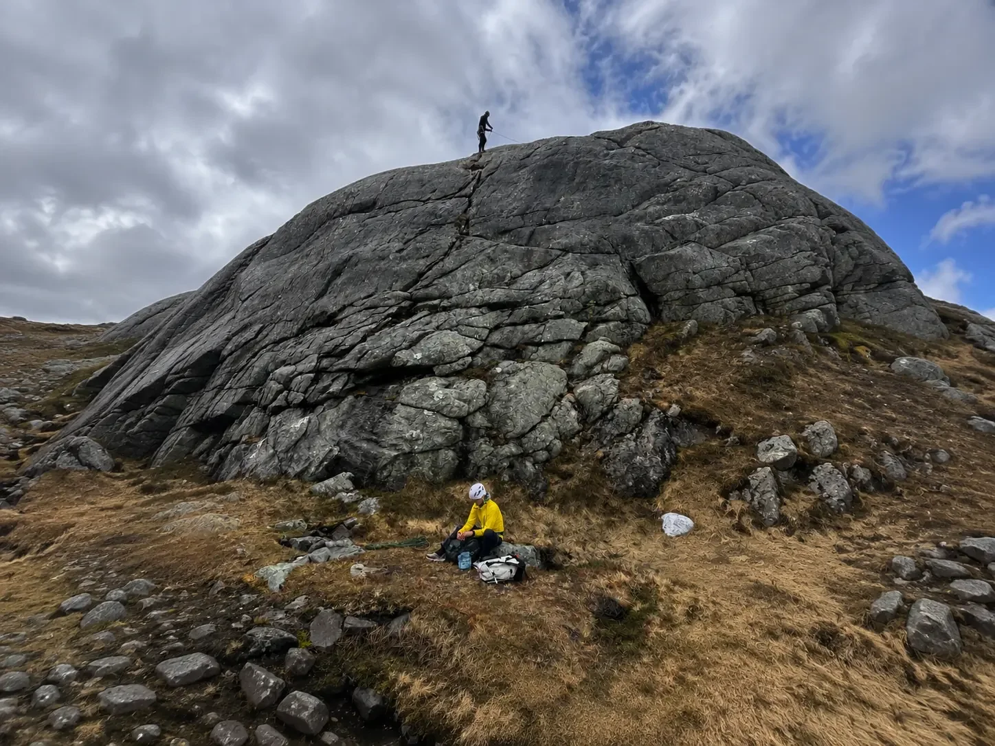 Hiker in yellow jacket and helmet resting on a grassy hillside with rocky terrain, while another hiker stands on a large boulder above, under a cloudy sky.