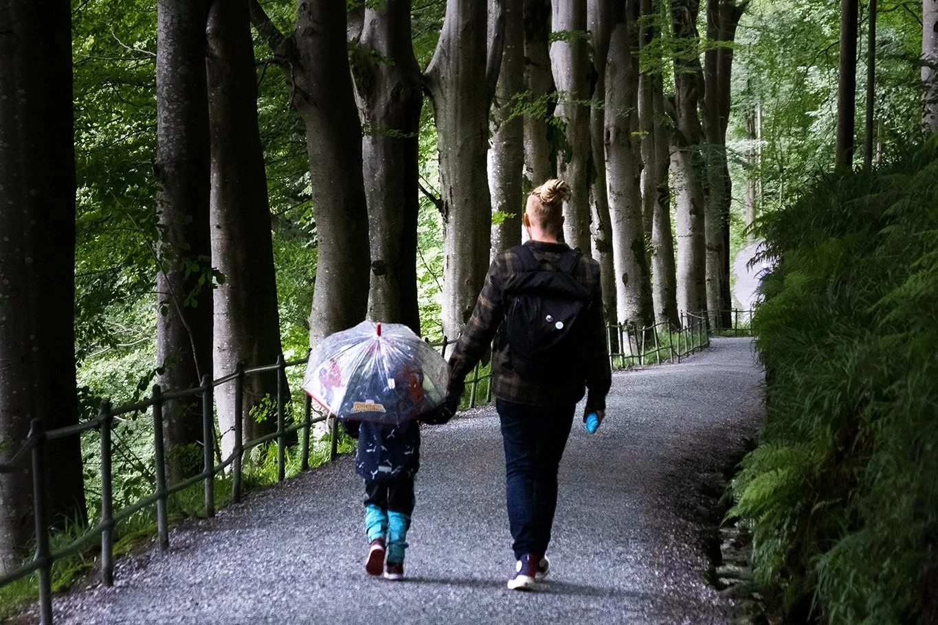 A woman and a child walking along a gravel path through a forested area with tall trees and green foliage, the child holding a clear umbrella.