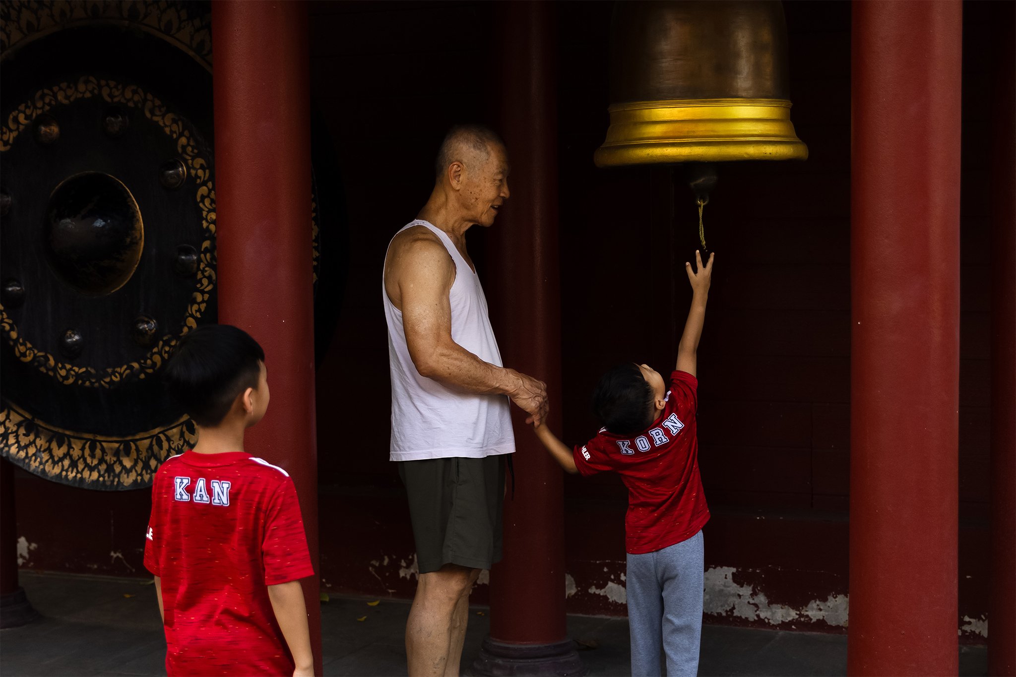 An elderly man and three children, two of whom are wearing red jerseys with the word 'KORN' on the back, are near a large bronze bell in a temple or similar setting. The man is smiling, and one child is reaching up to the bell.