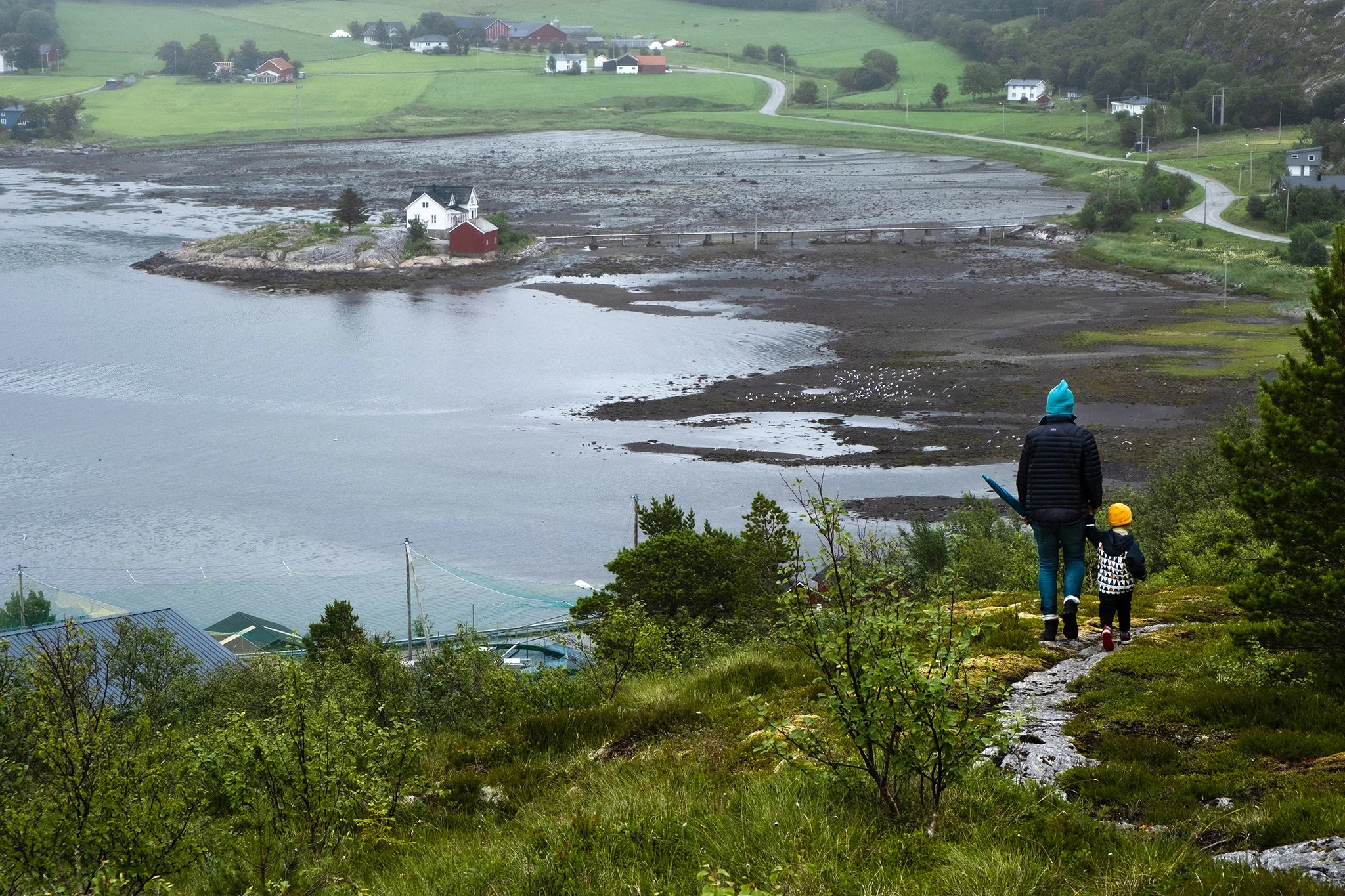 A woman and a child walk down a grassy hill with green foliage, overlooking a coastal area with a bridge, houses, and a body of water. The scene appears to be on a cloudy, cool day.