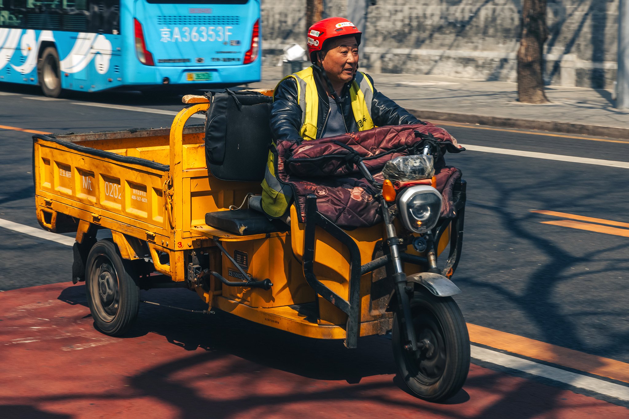 A man riding a yellow three-wheeled motorized vehicle carrying bags and belongings, on a city street with a blue bus in the background.