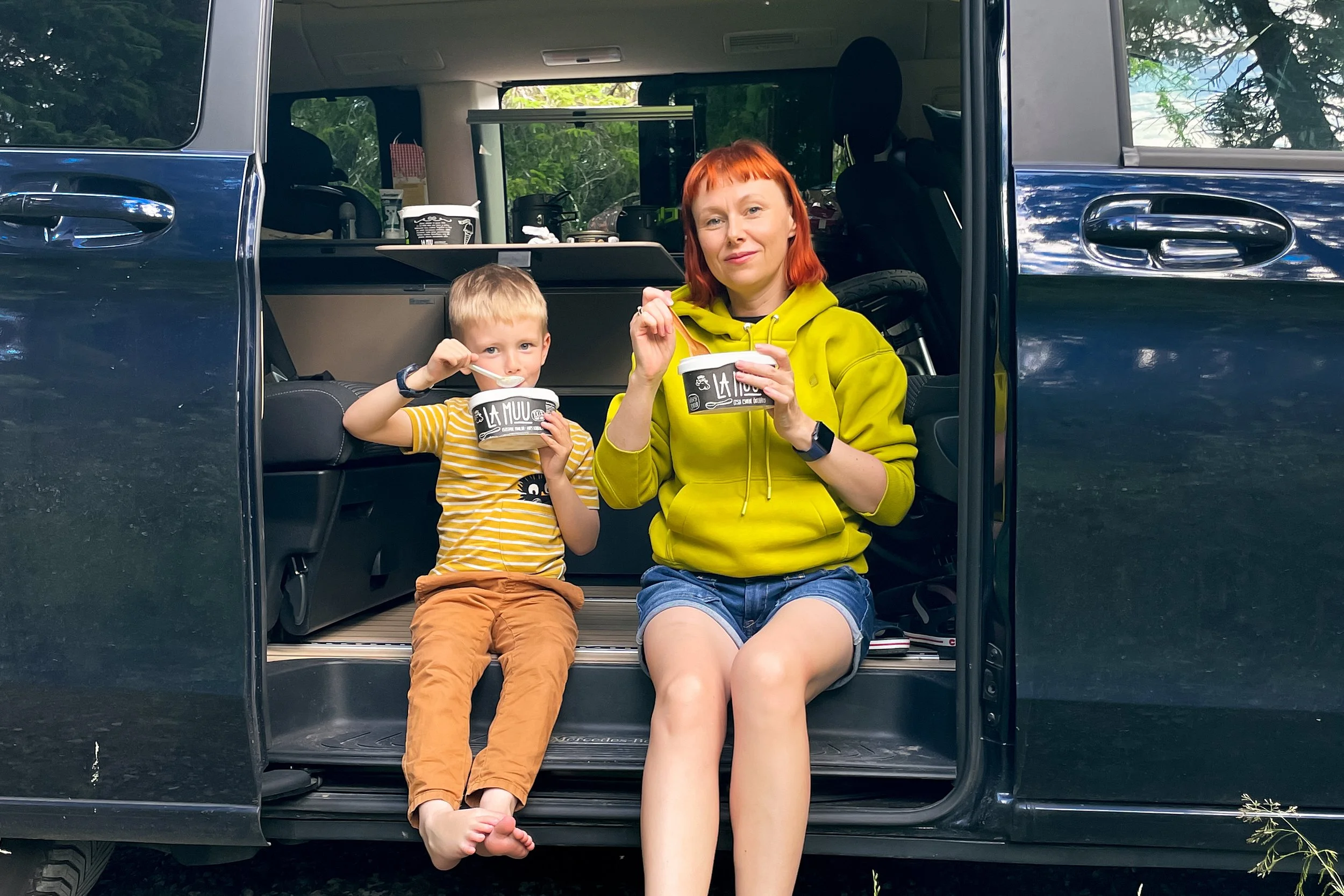 A woman with red hair and a young boy with blond hair sitting in the open back of a black SUV, eating ice cream. The woman is wearing a yellow hoodie and shorts, while the boy is in a yellow striped shirt and brown pants.
