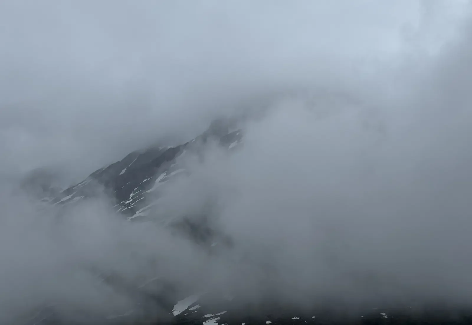 Cloud-covered mountain peak with patches of snow, obscured by thick gray clouds.