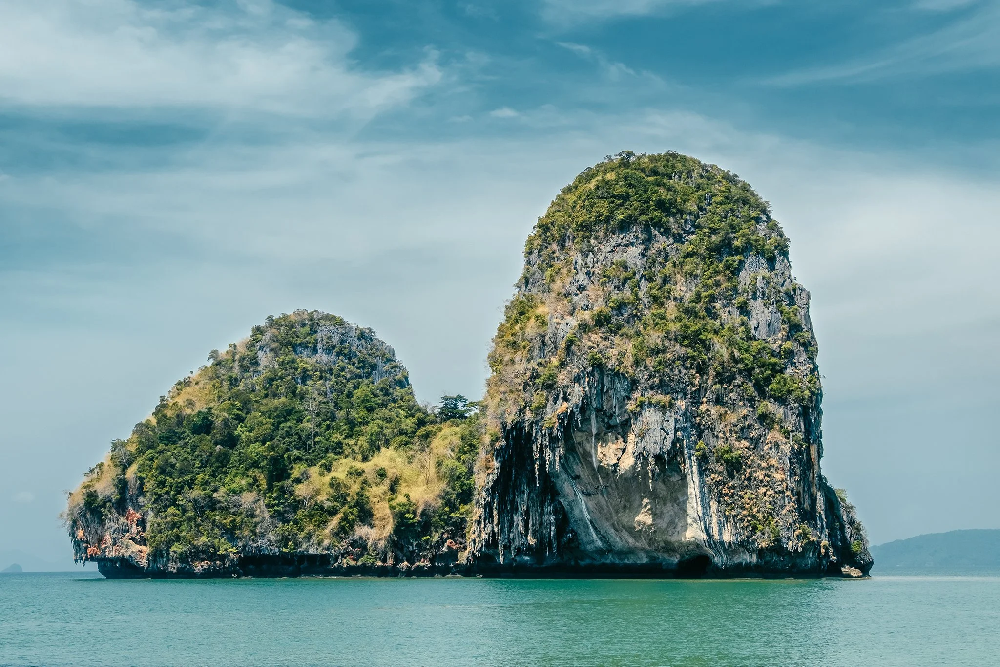 A large limestone island with lush greenery on top, surrounded by turquoise water and a blue sky with some clouds.