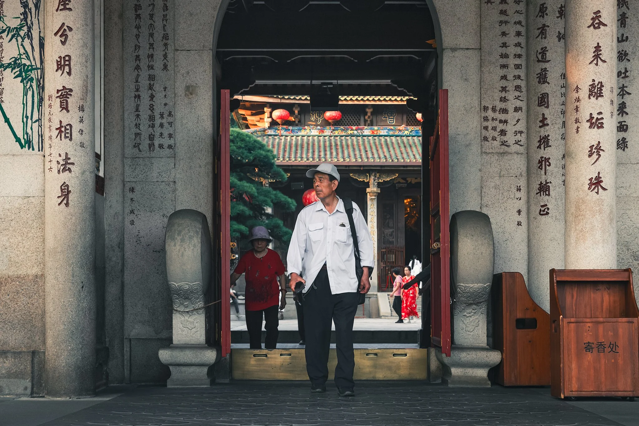 People walking through the entrance of a traditional Asian temple with Chinese characters on stone pillars and red lanterns hanging overhead.