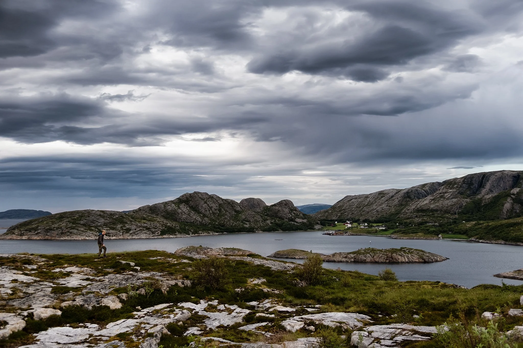 A person walking along rocky terrain near a body of water, with mountainous landscape and houses in the background under dark, cloudy sky.