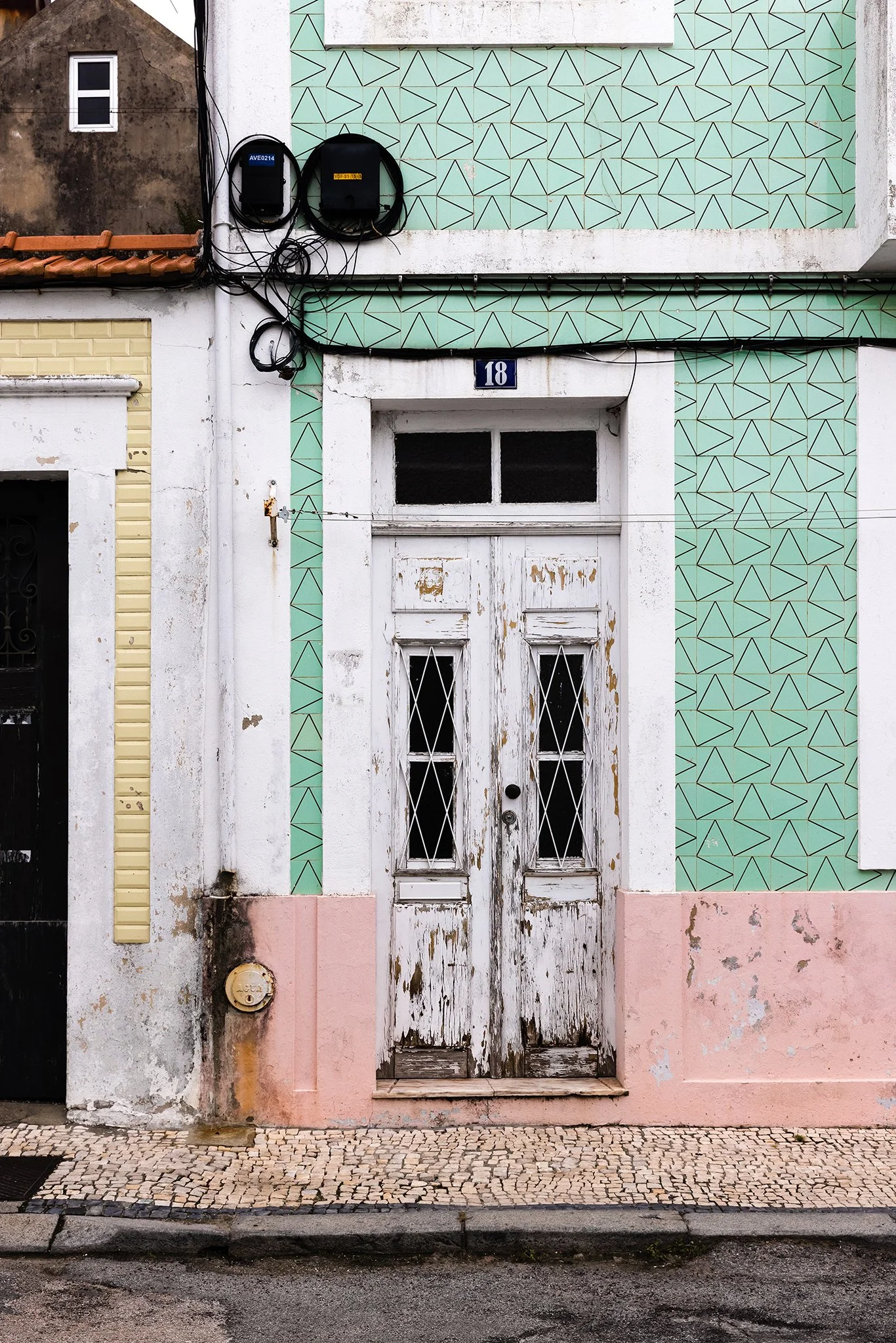 An old, weathered double door with peeling white paint on a building facade. The building features a green wall with a geometric pattern, a pink lower section, and a small blue number 18 plaque above the door. The street in front has cobblestone pavement.