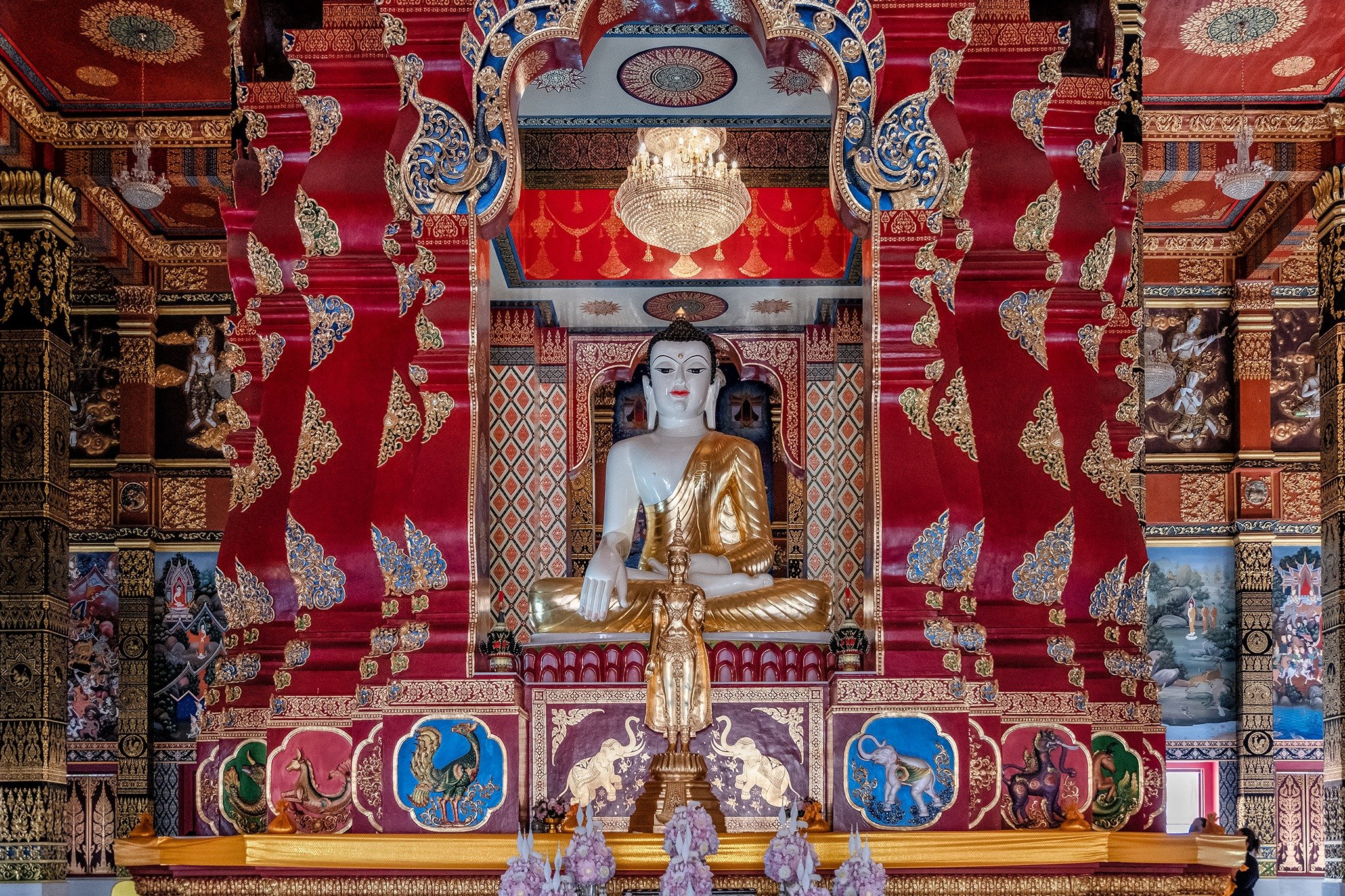Interior of a Buddhist temple with a large seated Buddha statue in the center, surrounded by ornate gold and red decorations, intricate carvings, and a chandelier hanging from the ceiling.