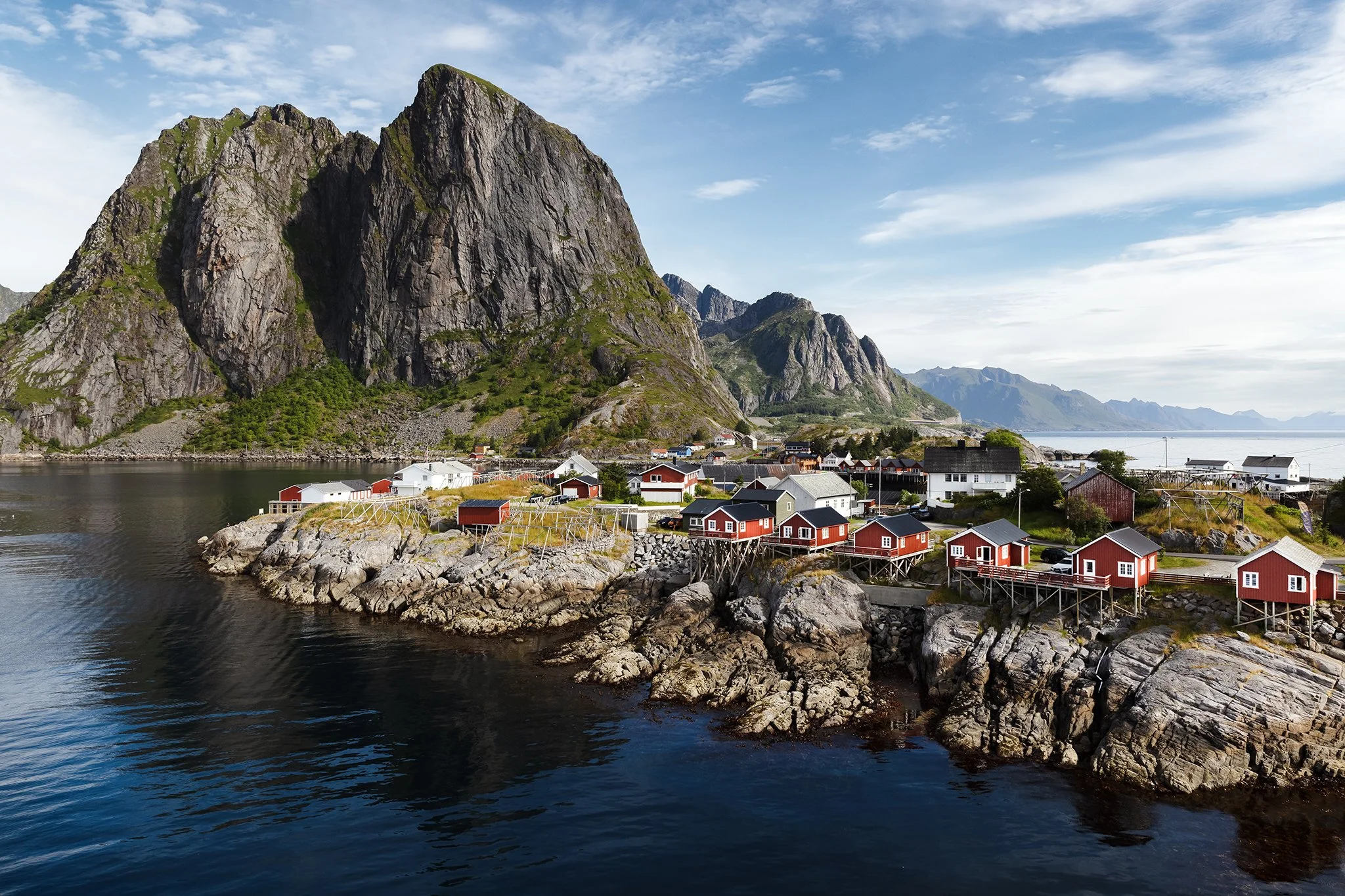 Small red and black houses beside a body of water with rocky shore, large mountain with green patches behind, and a partly cloudy blue sky.