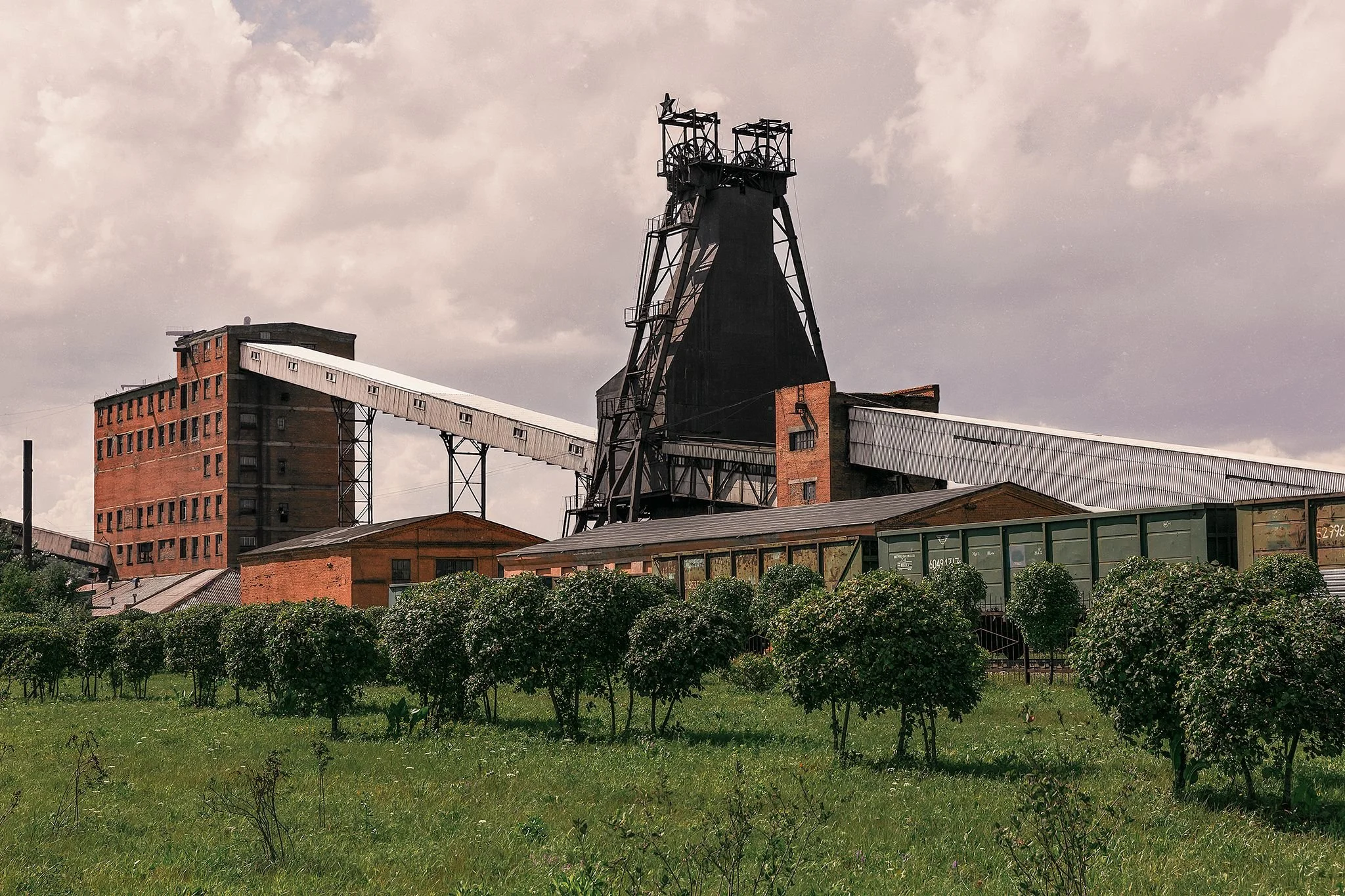 An industrial building with a tall black metal tower, brick and metal structures, and a conveyor belt extending from the building, set against a cloudy sky with a foreground of rectangular green bushes and a lawn.