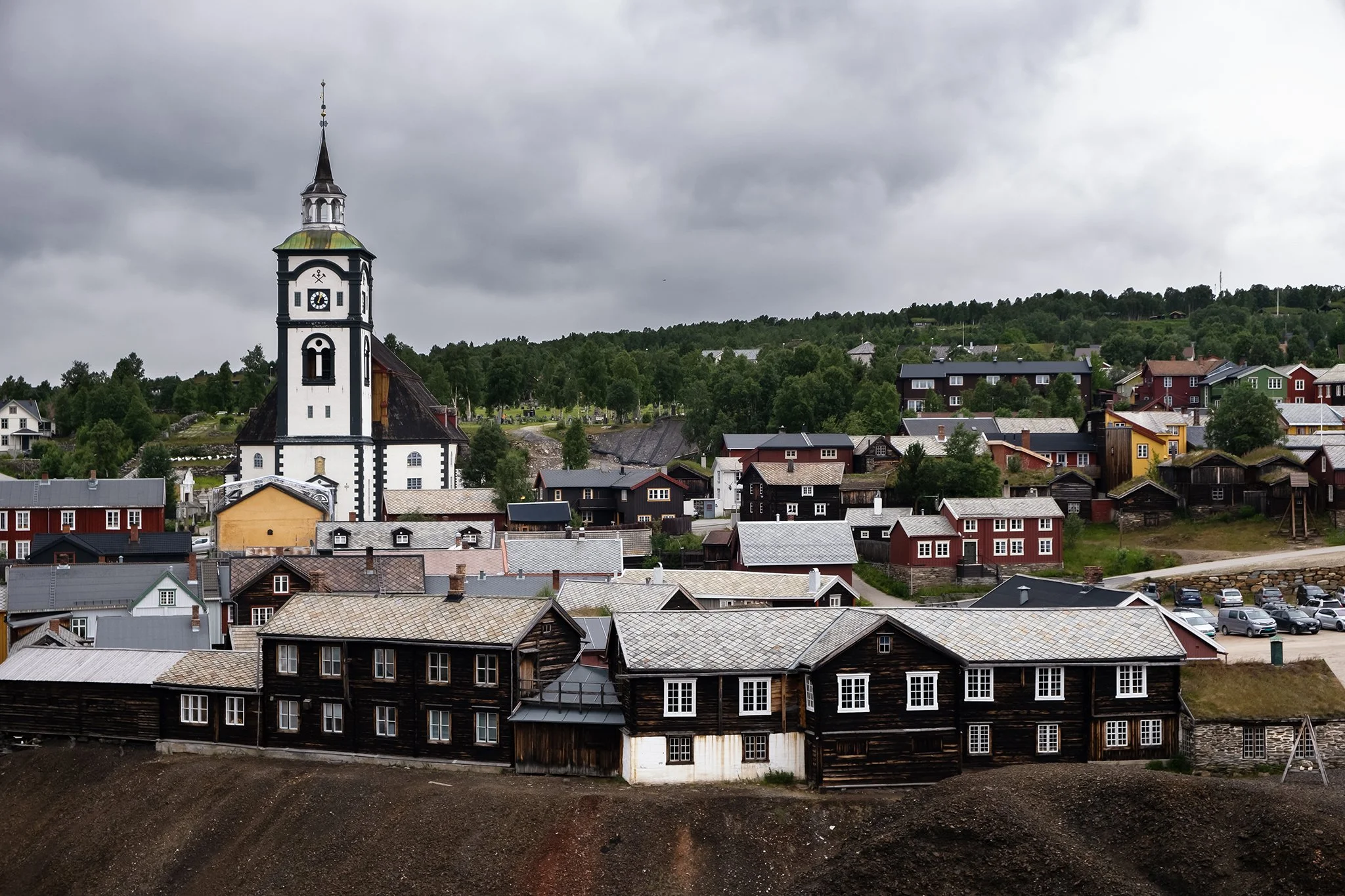A small town with a prominent white church featuring a tall steeple, surrounded by scattered houses with colorful and wooden exteriors, and green trees on a cloudy day.