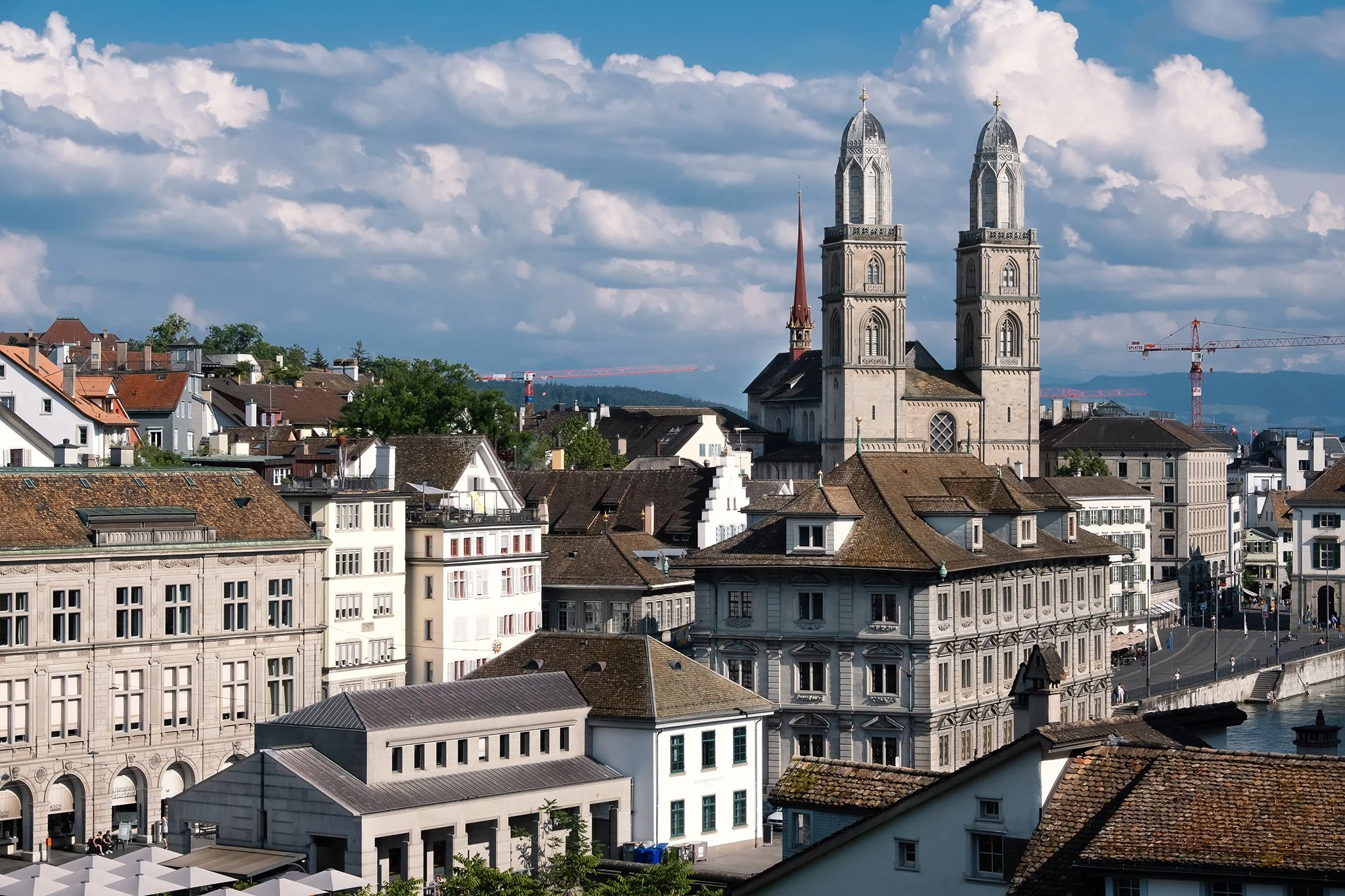 Cityscape featuring historic buildings with two prominent church towers, surrounded by modern structures and construction cranes, under a partly cloudy sky.
