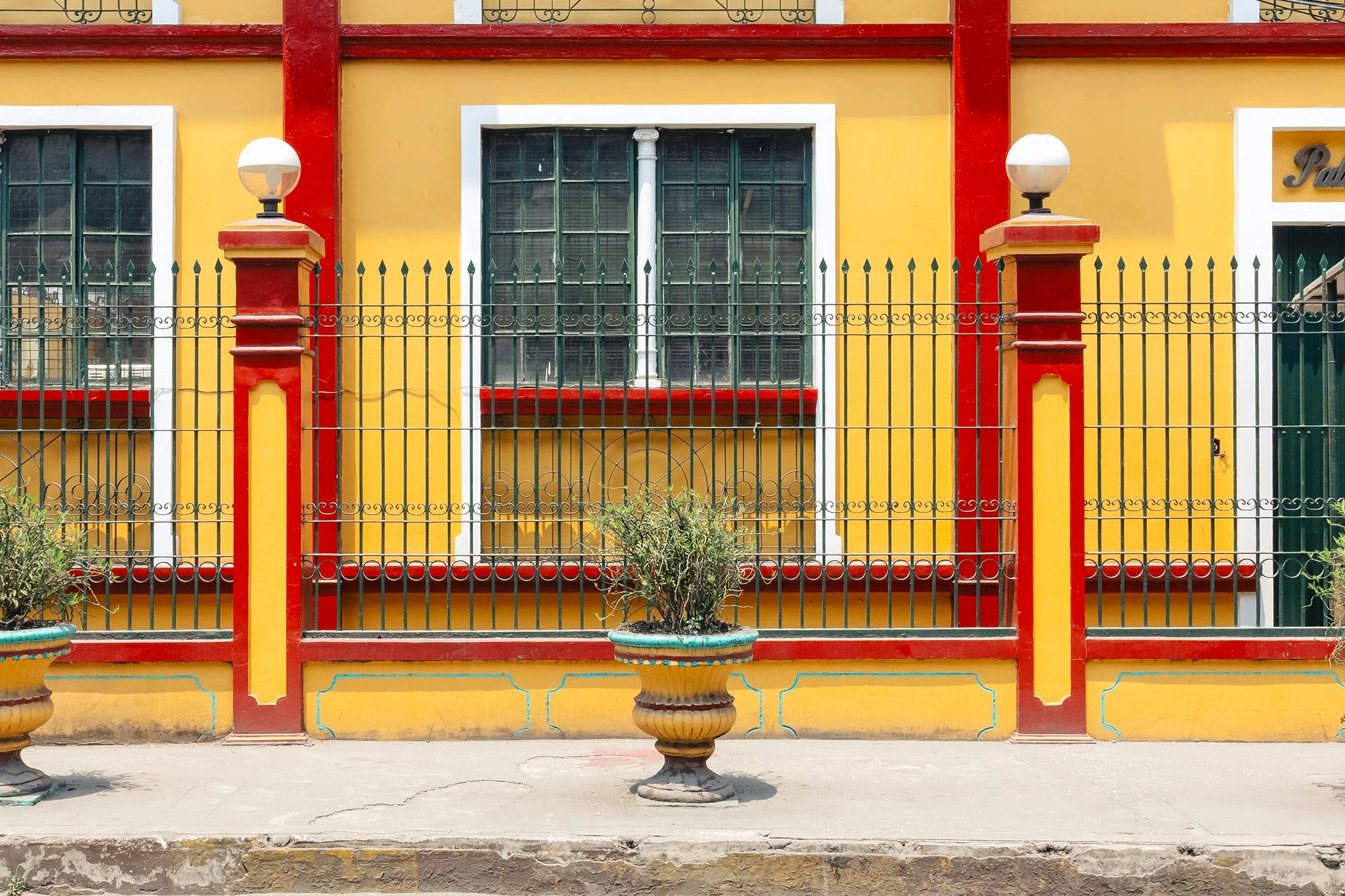 Bright yellow building with red trim and white-bordered windows, surrounded by a black wrought iron fence with decorative elements, and topped with two lamp posts. Small potted plants are in front.