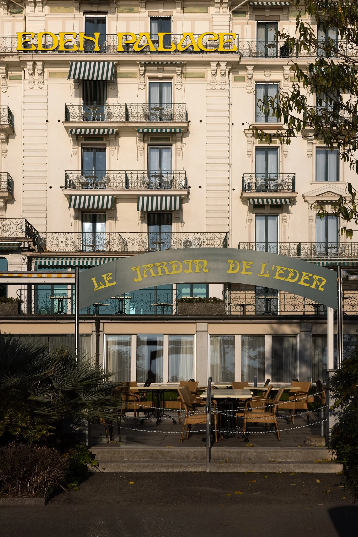 The image shows the front of a hotel building named Eden Palace, with multiple floors and balconies with wrought iron railings. Some balconies have striped green and white awnings. There is an entrance with a sign that reads 'Le Jardin de Leden,' and outdoor seating with tables and chairs in front of the building, surrounded by plants.