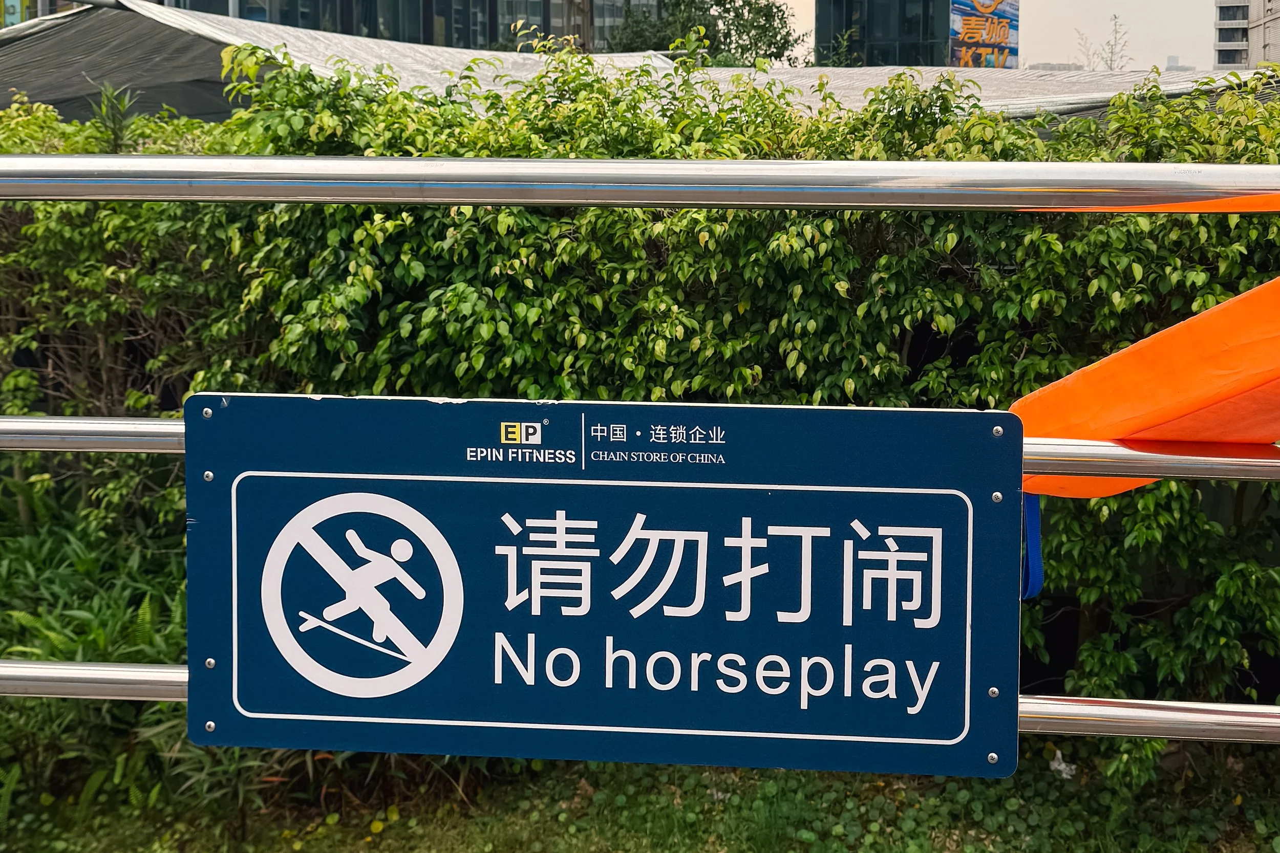 Blue and white sign with a no-horseplay symbol and Chinese characters, located near green bushes, with a background of a metal railing, trees, and buildings.