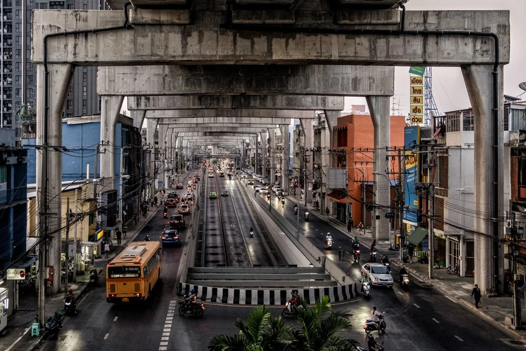 City street view taken from an elevated metro track, showing traffic, motorcycles, and buildings on both sides under cloudy skies.