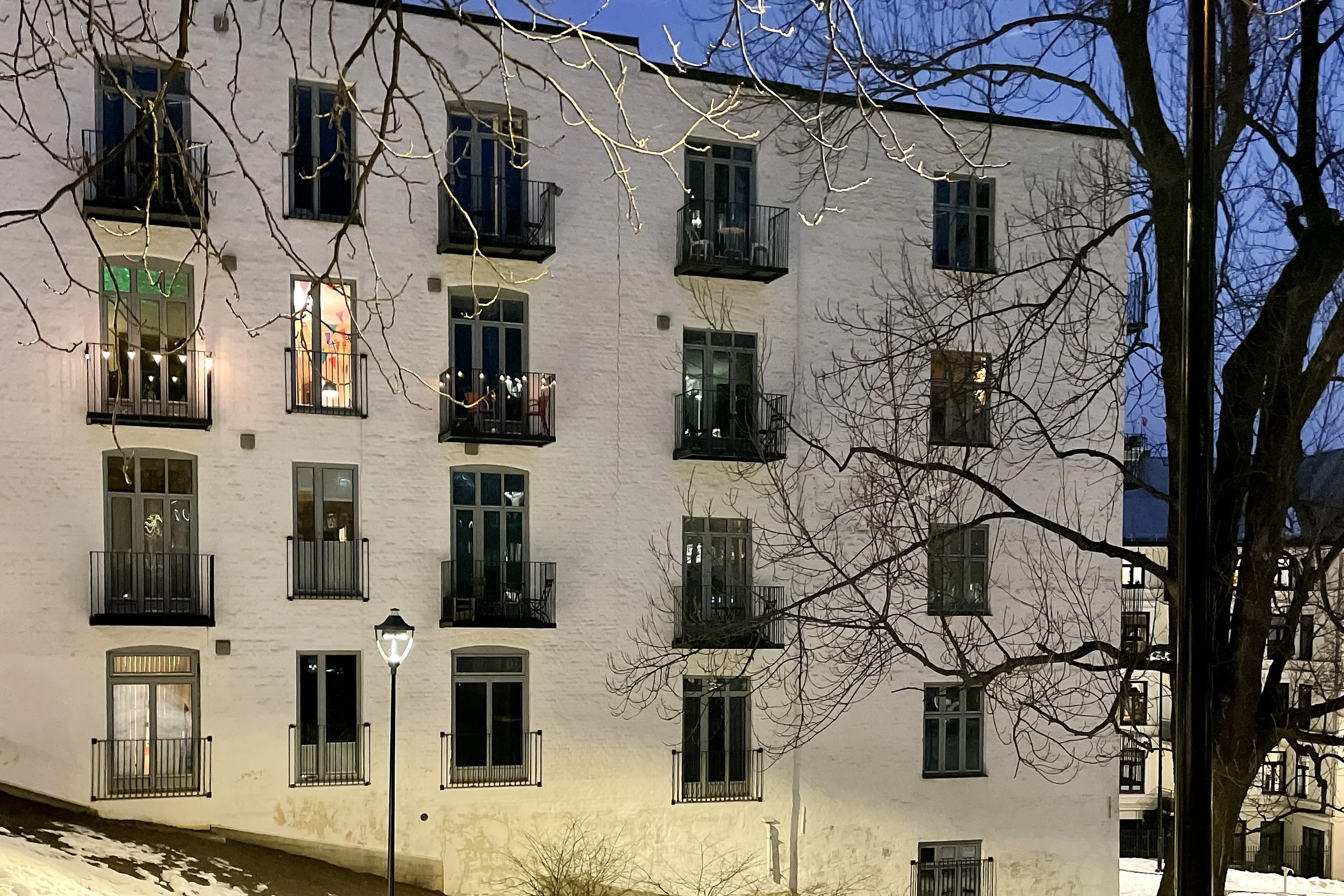 A white multi-story building with several balconies and windows, some of which are illuminated. The building is surrounded by leafless trees, and streetlights line the sidewalk in the foreground. It appears to be evening or nighttime, with a dark sky overhead.
