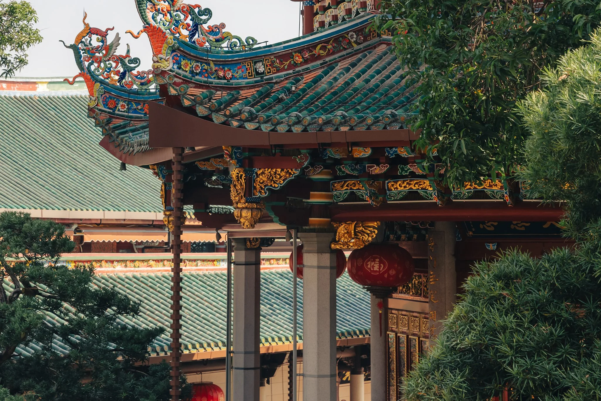 Close-up of a traditional Chinese temple roof with ornate, colorful decorations, including dragons and floral designs, partly obscured by green trees.