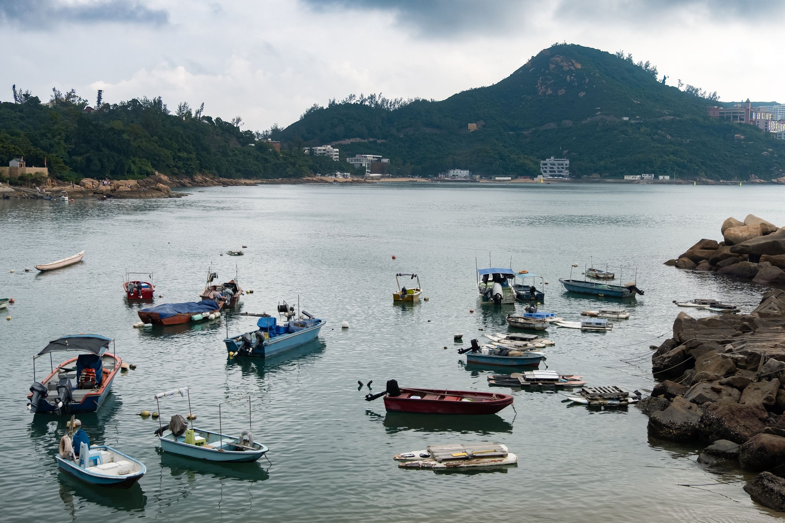 A harbor with several small boats floating on calm water, some tied to rocks on the rocky shoreline in the foreground, with green hills and buildings in the background under cloudy skies.