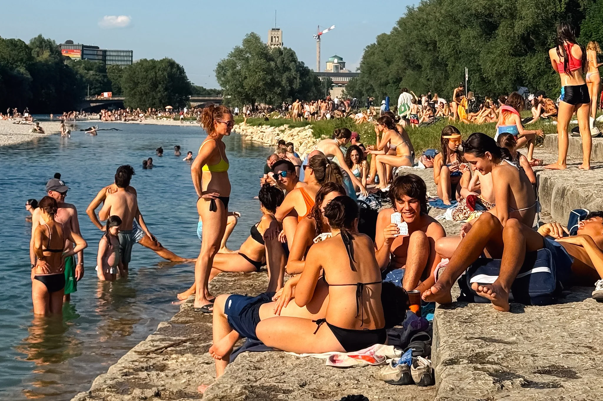 Crowded riverside beach with many people sunbathing, sitting, and swimming on a sunny day, surrounded by green trees and city buildings in the background.