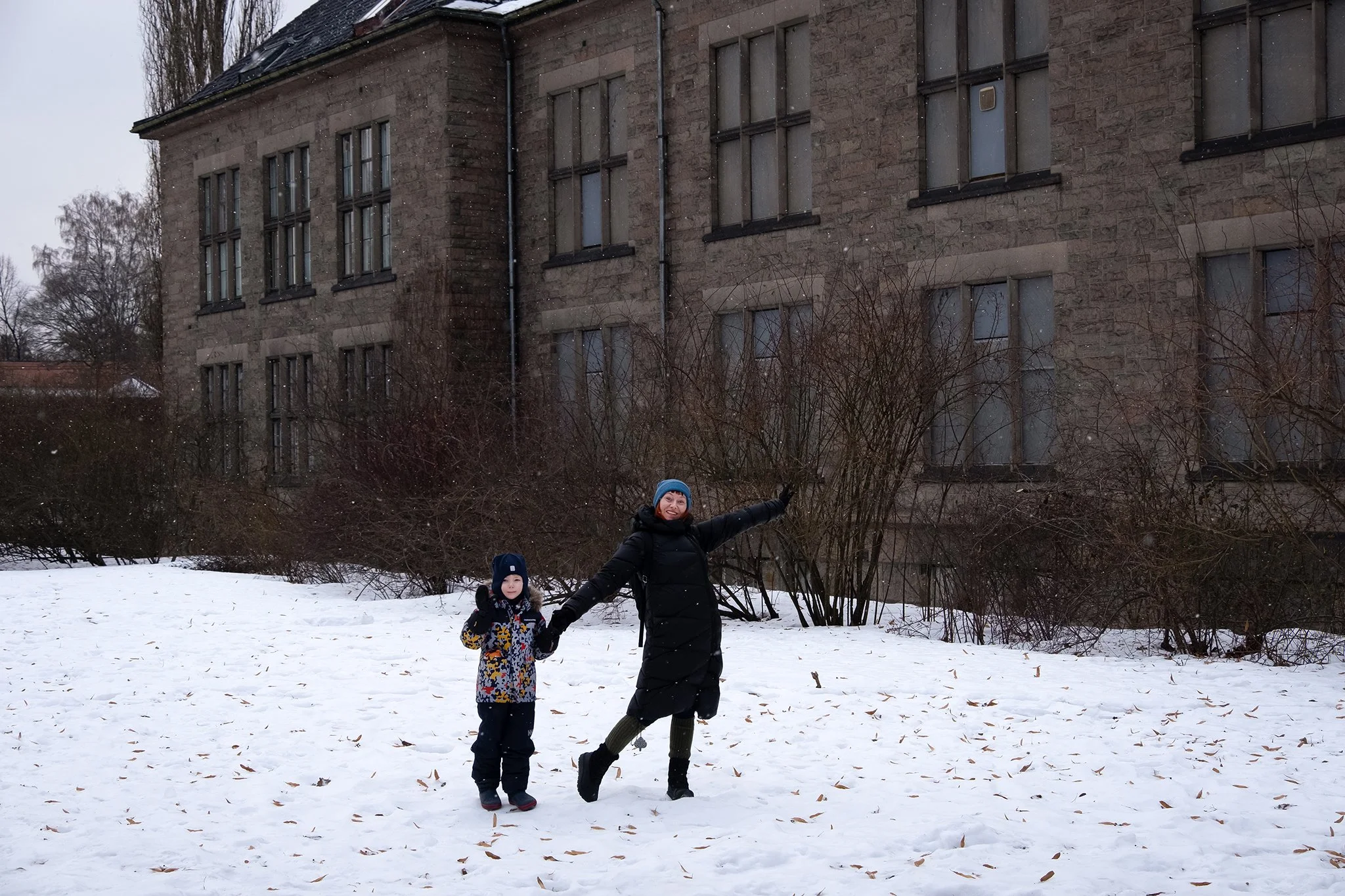A woman and a young girl stand in a snowy outdoor area, smiling and holding hands. The woman is wearing a black winter coat, green pants, and a blue hat, while the girl is dressed in a patterned winter coat and dark pants. Behind them is a large brick building with multiple windows and leafless bushes, with snow falling.
