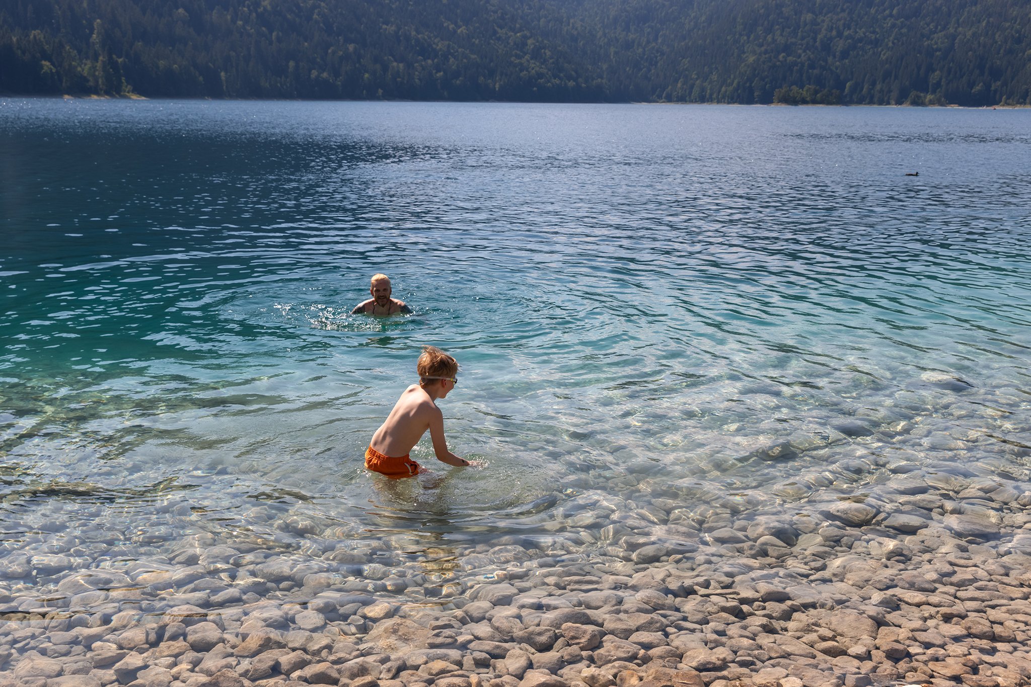 Two children playing in clear water near a rocky shore, with a man swimming behind them and a large lake with a forested mountain in the background.