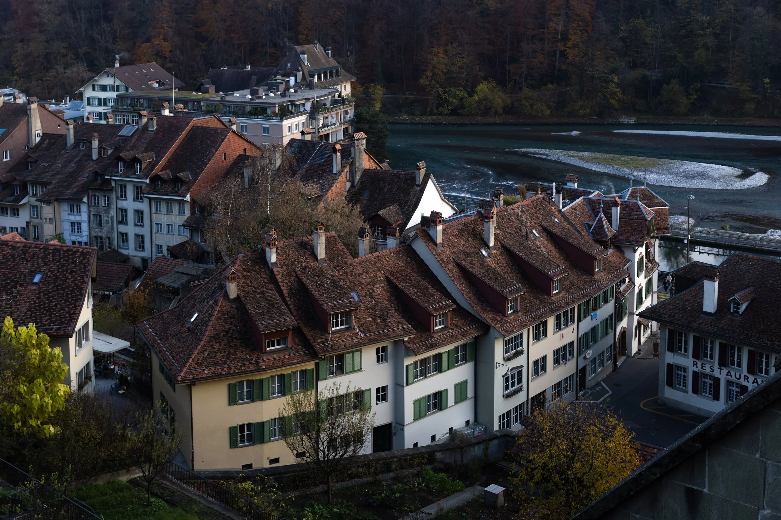 Aerial view of European-style buildings with red-tiled roofs next to a river and wooded hills.