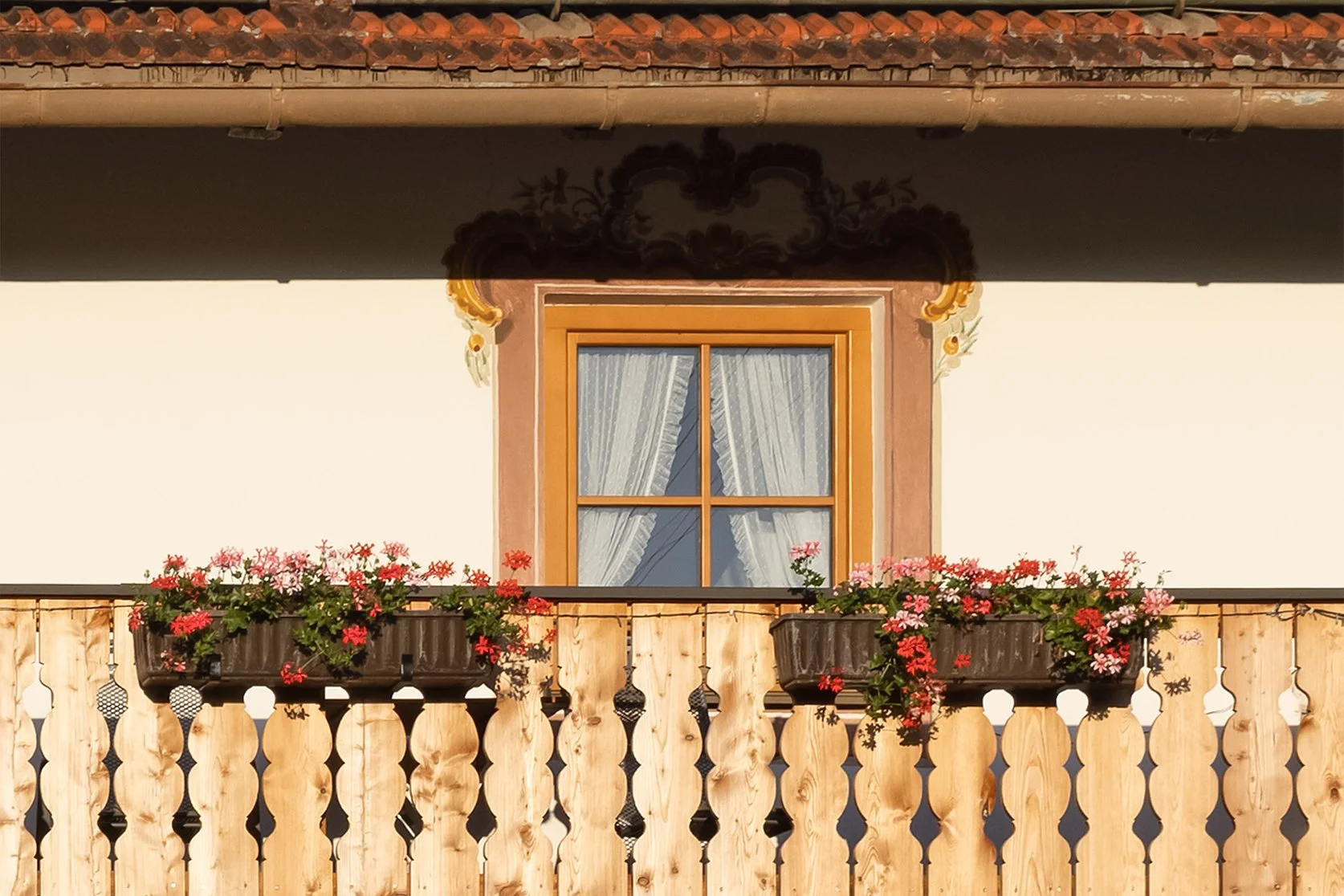 A house window with wooden frame and white curtains, surrounded by decorative brown trim, above a wooden balcony railing with flower boxes filled with red and pink flowers.