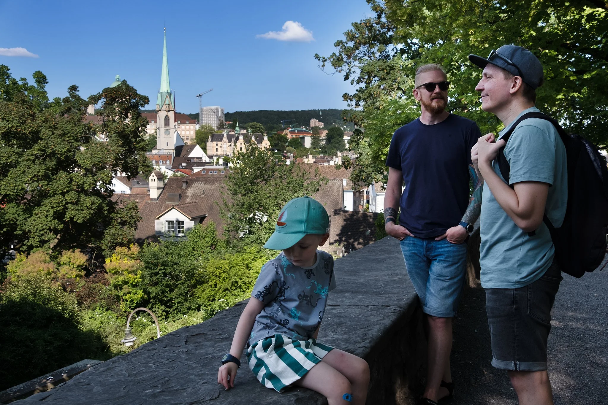 Two men and one child standing outdoors in a park with a cityscape in the background, including a tall church steeple, trees, and buildings. The man on the right is wearing a gray t-shirt, a black cap, and sunglasses, while the man on the left has a beard, glasses, and is dressed in a black t-shirt. The child is sitting on a stone ledge, wearing a gray shirt with a dinosaur print and striped shorts, looking down.