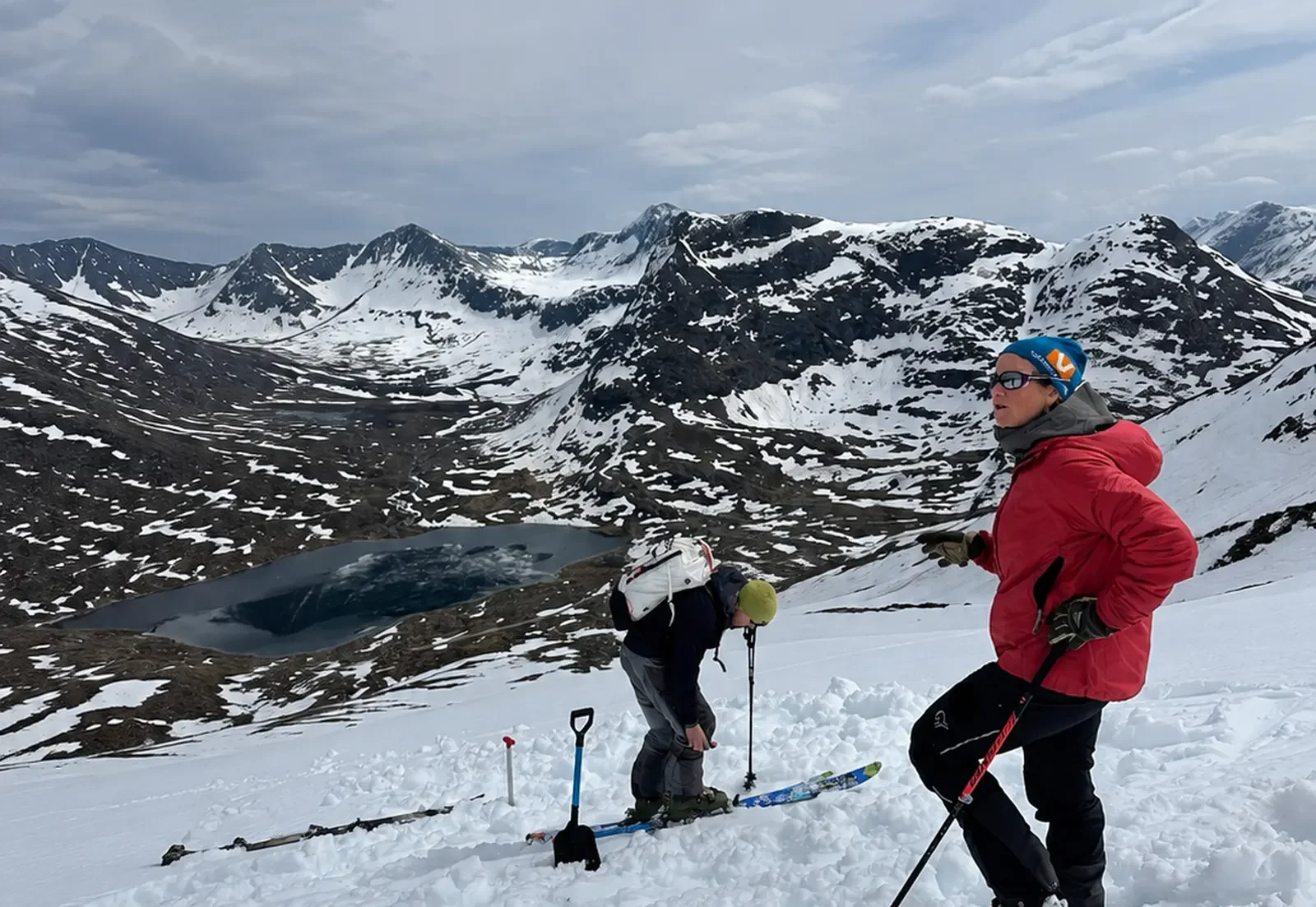 Two skiers in red and black gear standing on snowy mountain slope with a scenic view of snow-covered mountains, a lake, and cloudy sky in the background.