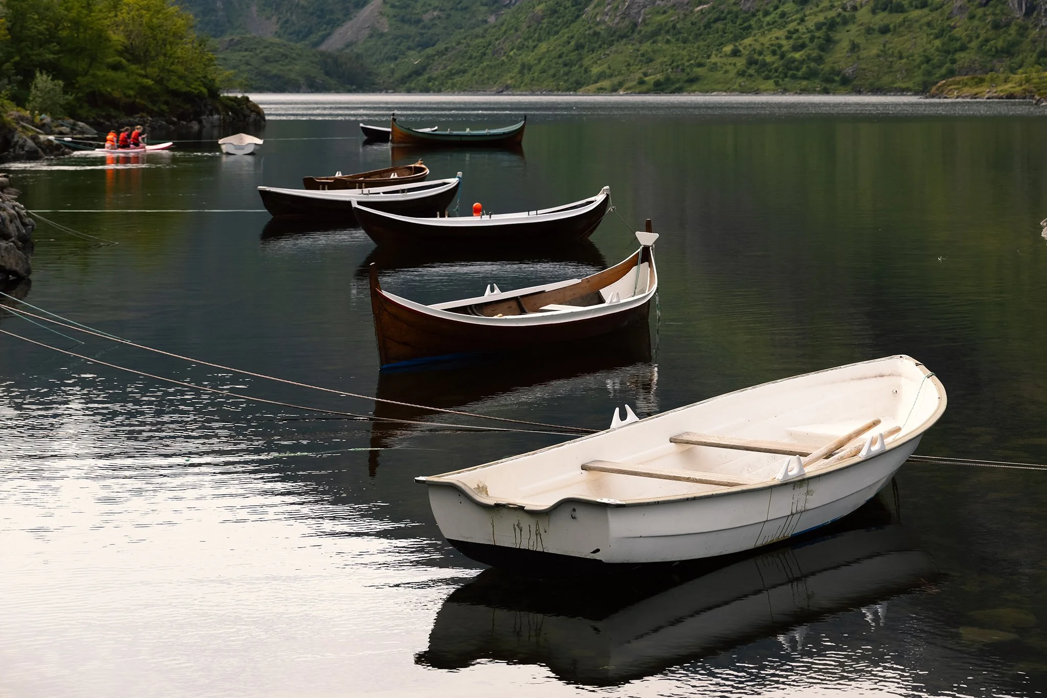Several small boats are docked on a calm lake with green hills and mountains in the background. Two people in red life jackets are on a small boat near rocks on the left side.