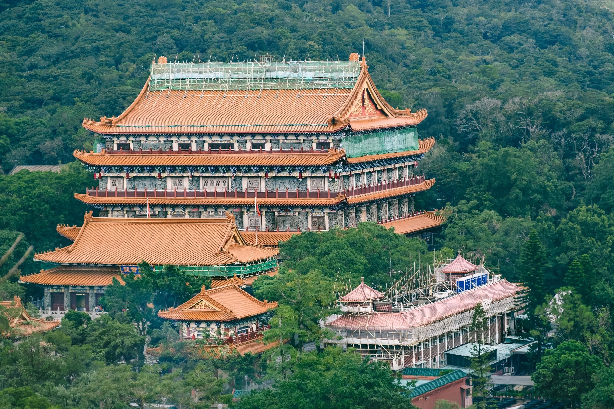 A traditional multi-tiered Chinese temple with orange tiled roofs and decorative architectural details, partially covered with green scaffolding and surrounded by lush green forest.