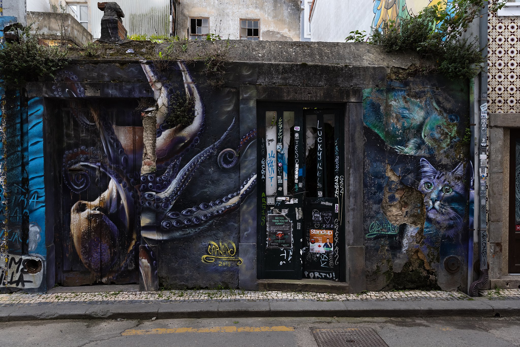 Street art mural of a purple octopus tentacle and a colorful cat face painted on a weathered building wall, surrounded by graffiti and stickers.