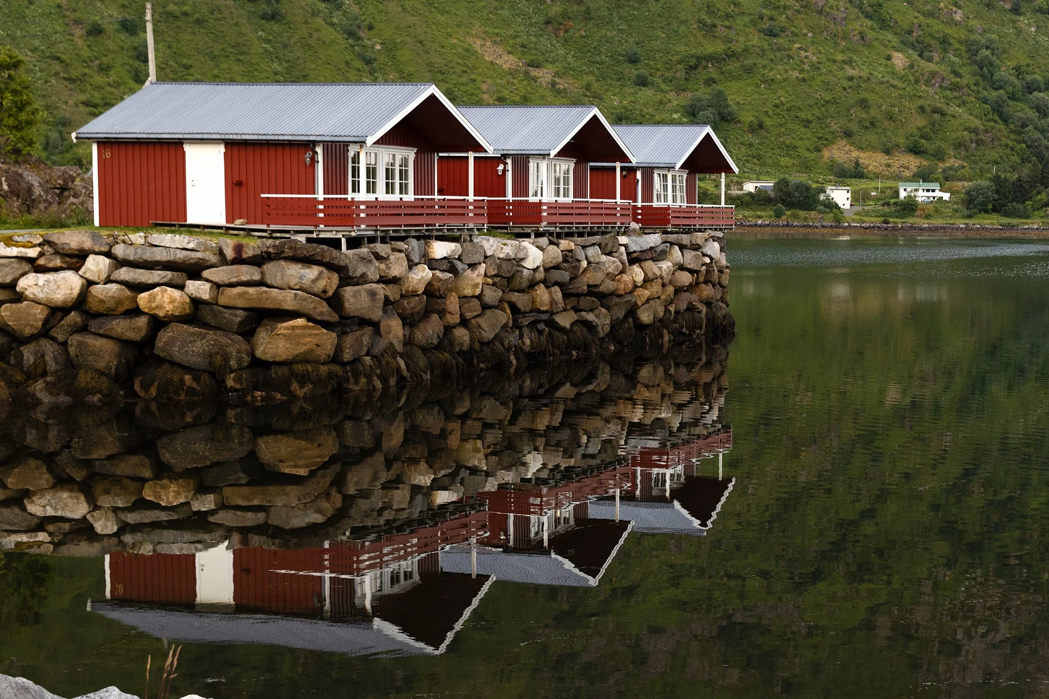 Red wooden cabins with white trim are built on a stone foundation along a lake with a reflection of the cabins in the water, and a lush green hillside in the background.