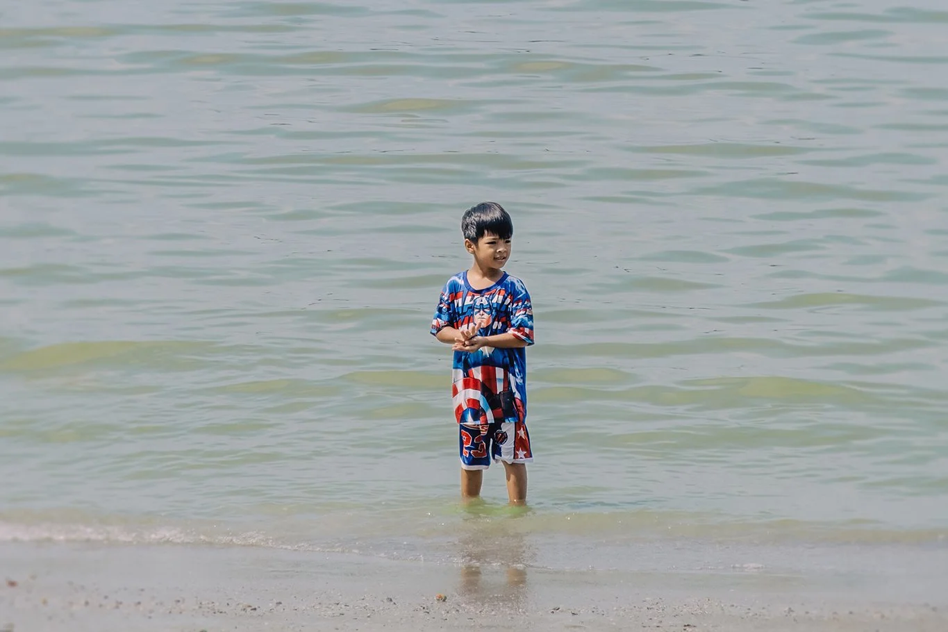 A young boy standing in shallow water at the beach, wearing a Captain America-themed swimsuit, smiling and looking to the side.