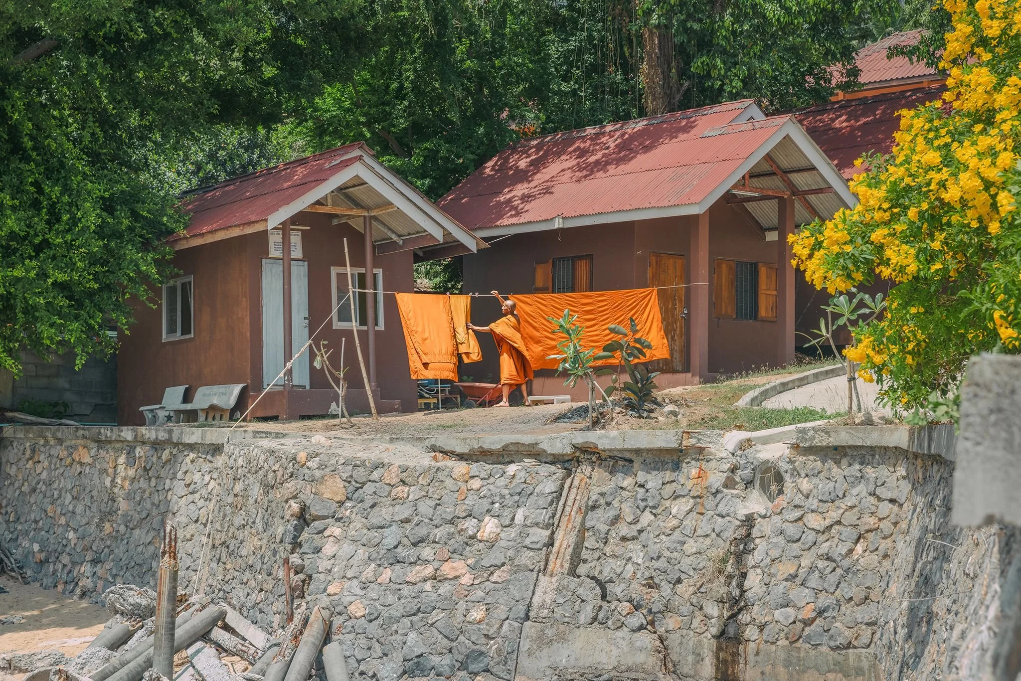 A woman in orange robes hanging laundry on a clothesline in a rural area with green trees and houses with red roofs.